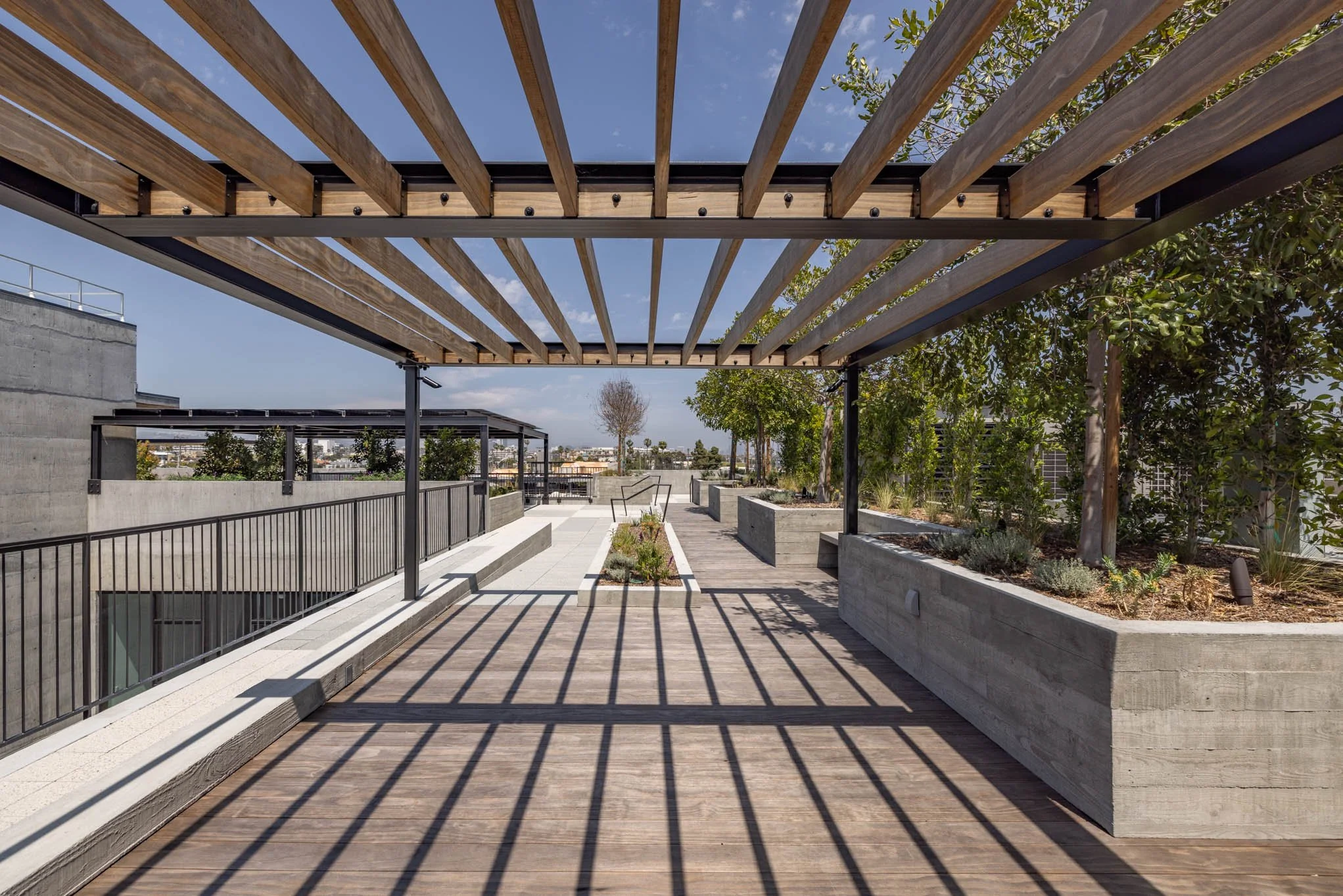 Rooftop terrace with wooden pergola, concrete planters with greenery, and city views, under a partly cloudy sky.