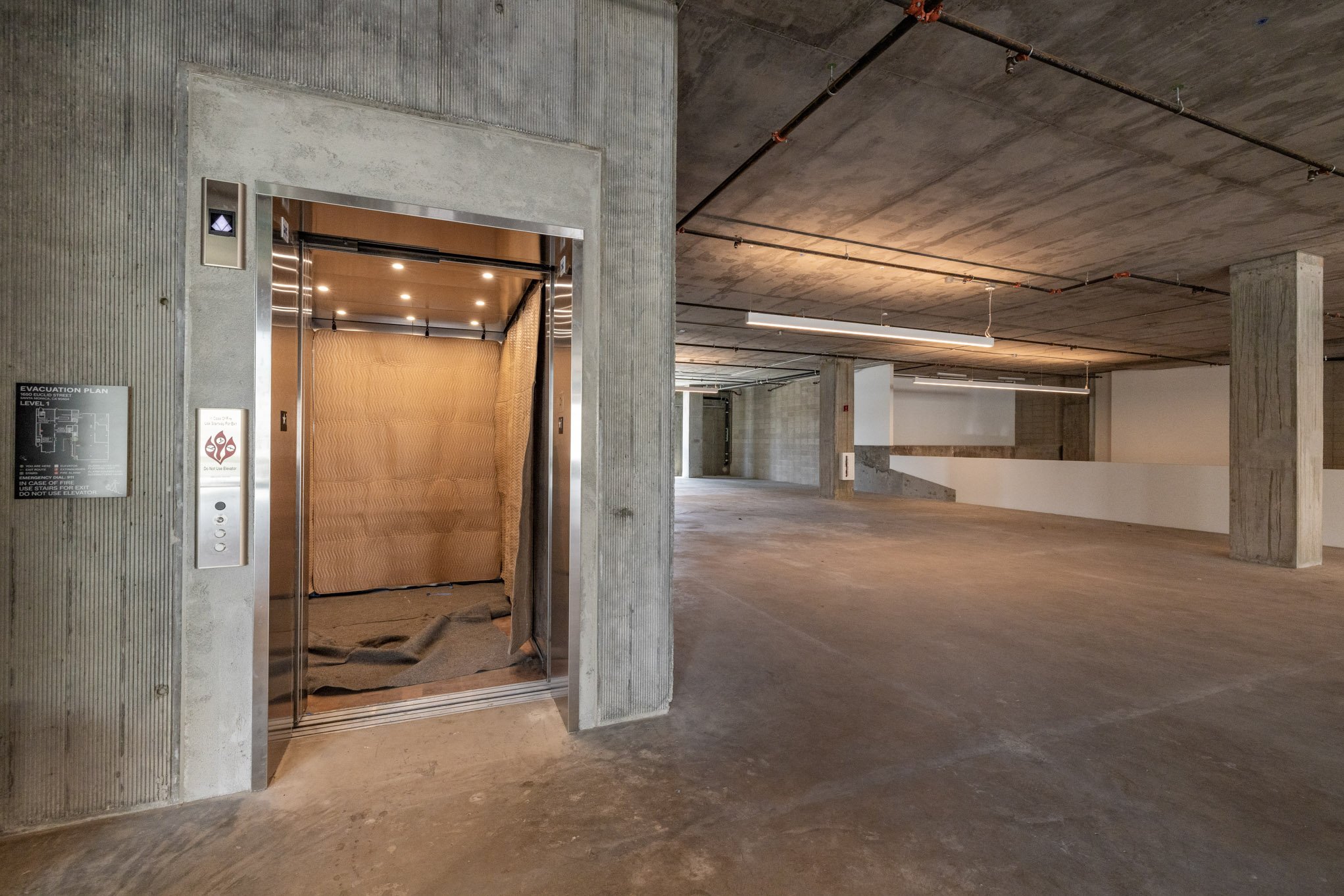 Empty underground parking garage with an elevator, concrete walls, floor, and ceiling, and pipe fixtures on ceiling.
