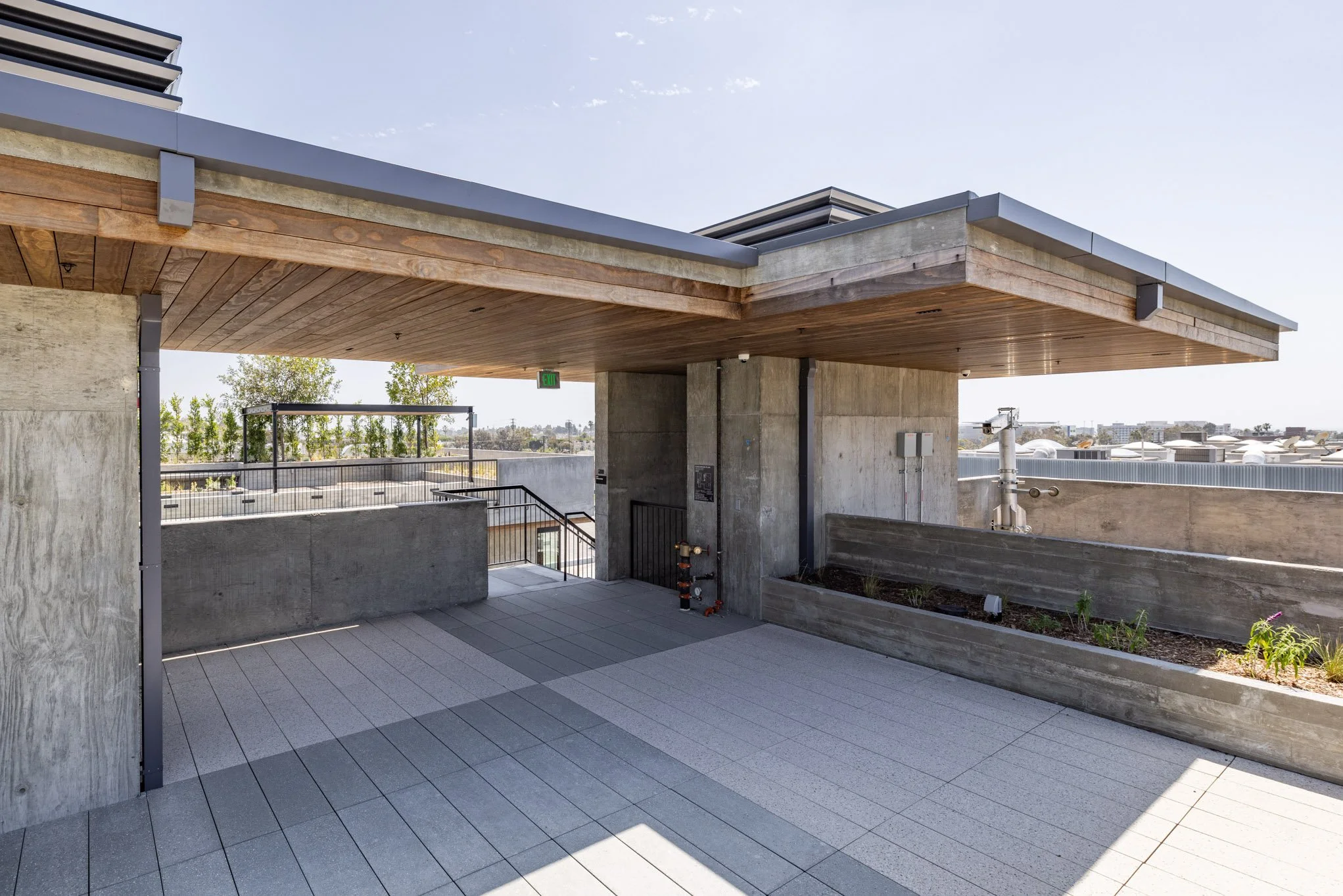 Rooftop terrace with concrete walls, wooden ceiling, planter box with plants, paved flooring, and cityscape in the background.