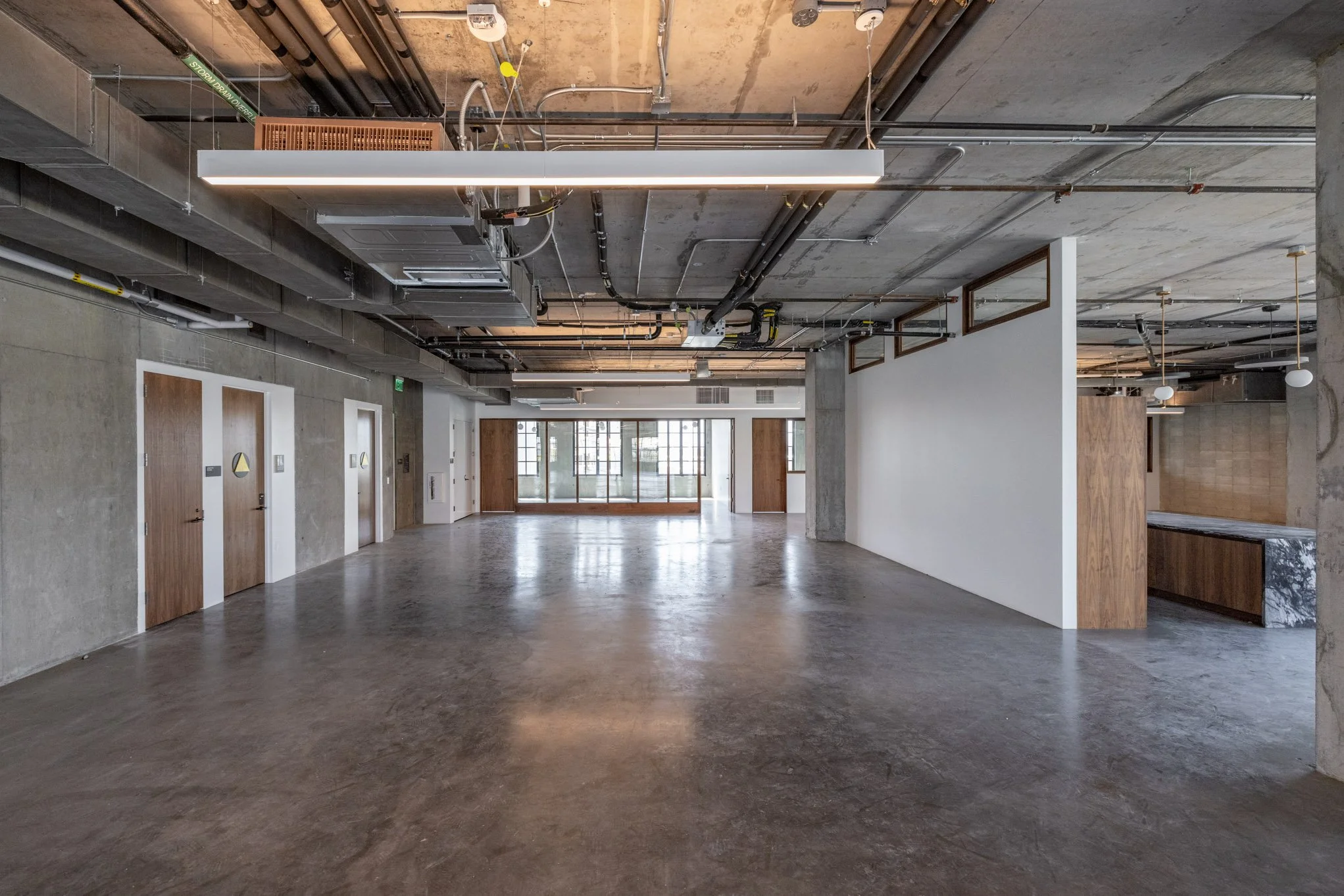Empty modern industrial-style interior space with concrete floors, exposed ceiling pipes, and wooden accents, including doors and framing, natural light coming through large window at the back.