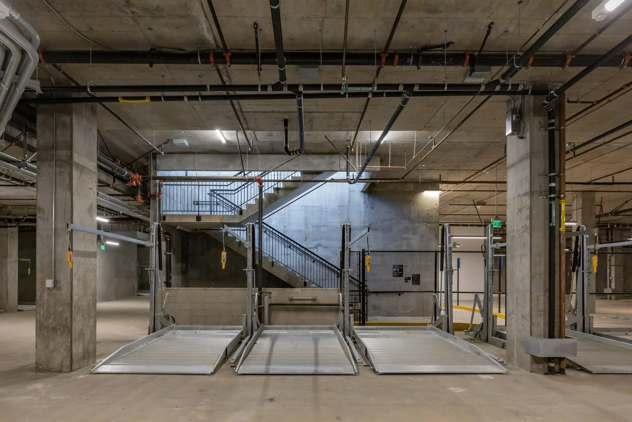 Underground parking garage with wheelchair accessible ramps, concrete pillars, staircase in the background, and industrial ceiling pipes.