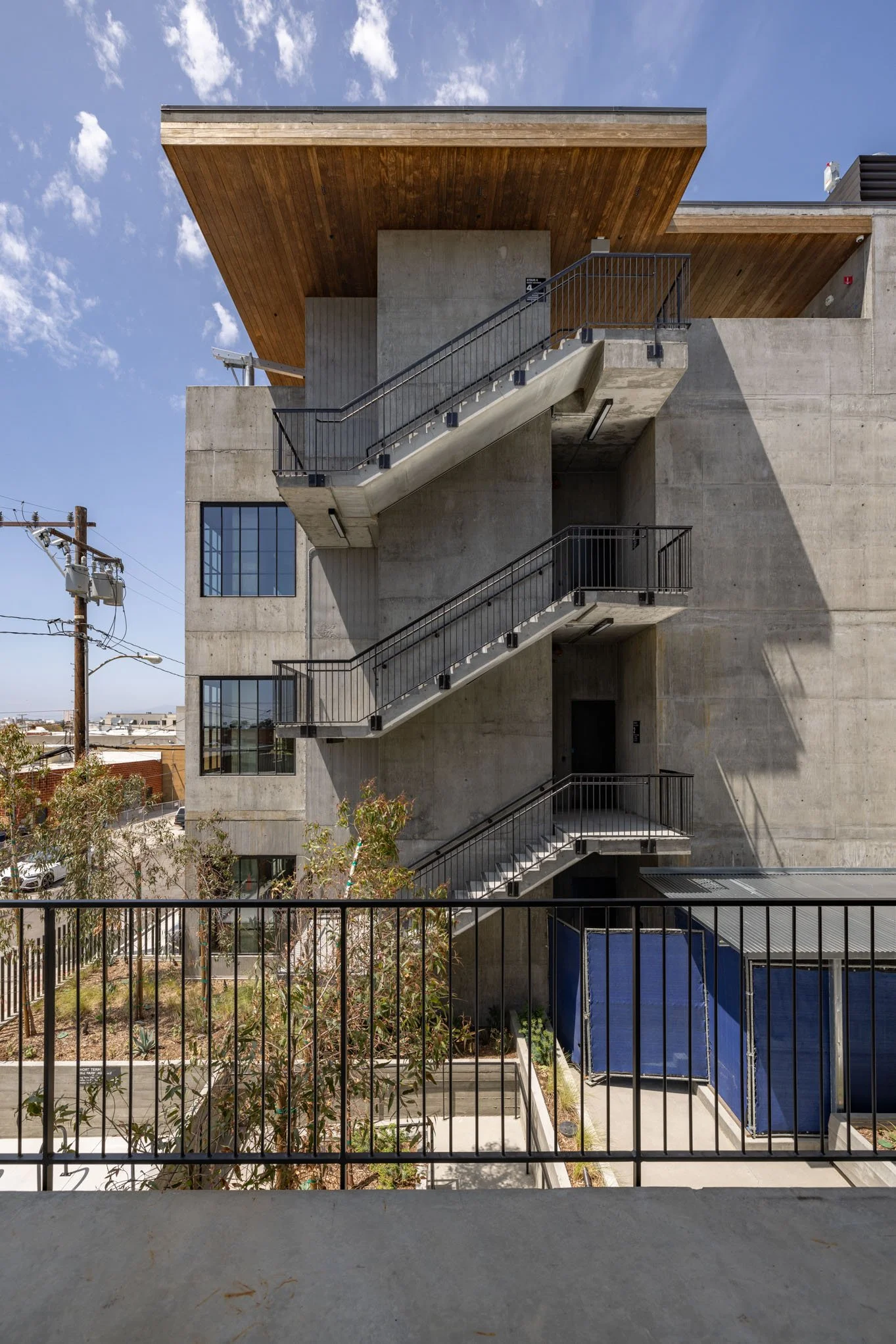 Modern multi-story concrete building with external metal staircases casting shadows on the wall, against a blue sky with a few clouds.