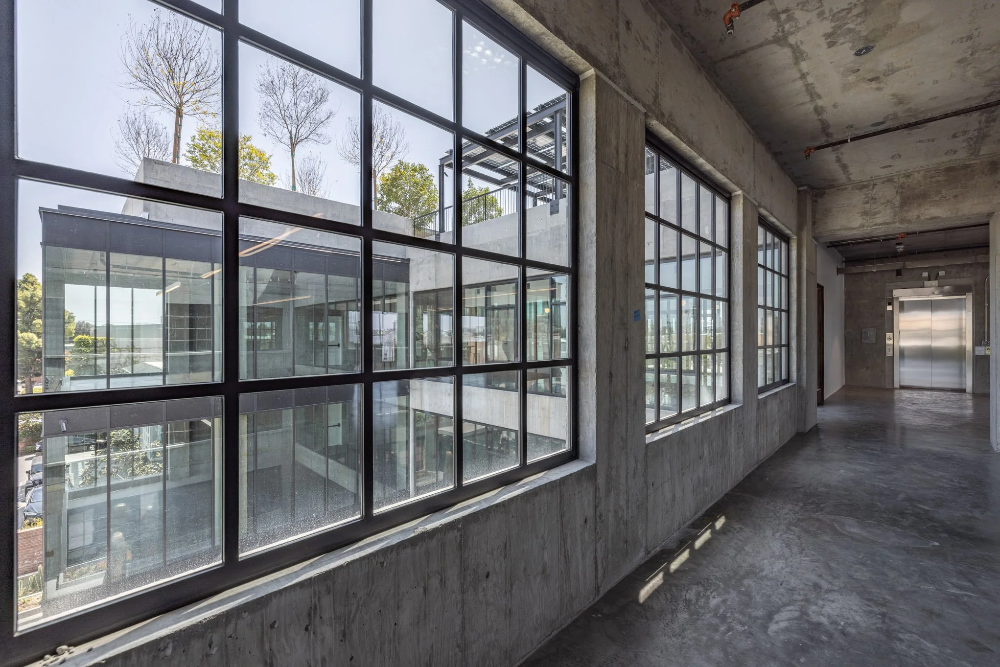 Interior view of a modern building under construction with large grid-style windows, unfinished concrete floors, and exposed concrete walls and ceiling.