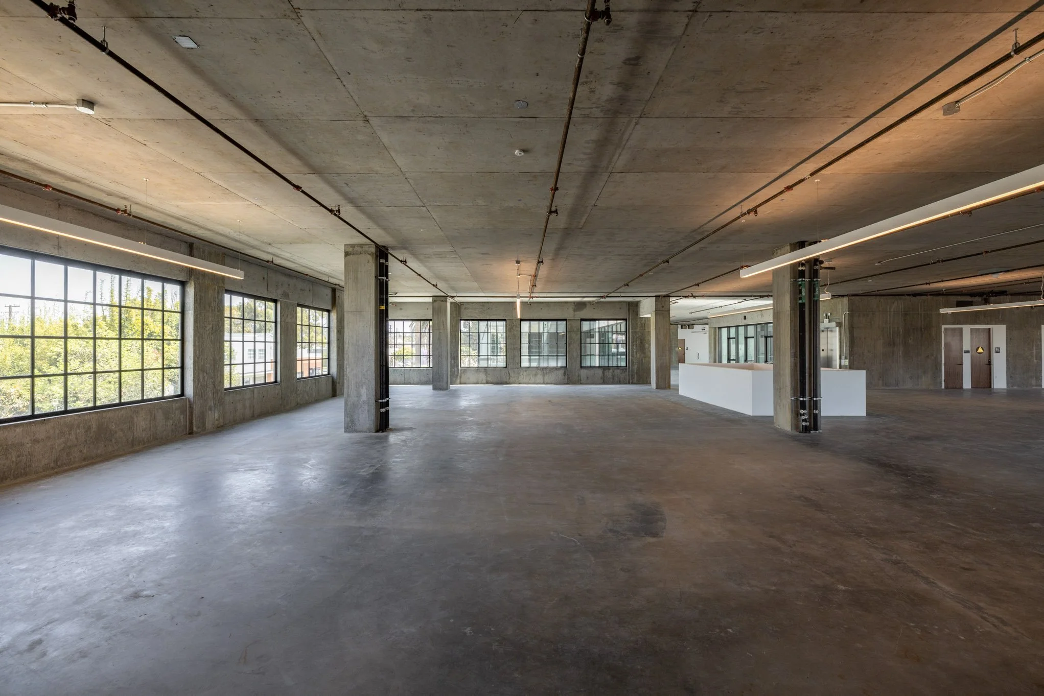 Empty industrial-style interior space with large windows, concrete floors, exposed ceiling, and minimal lighting fixtures.