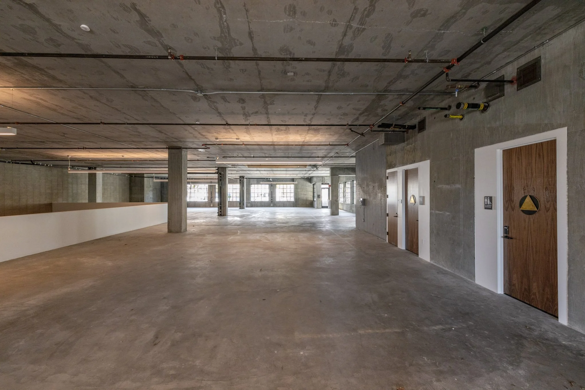 Empty industrial space with concrete floor, exposed pipes, and windows on the far wall.