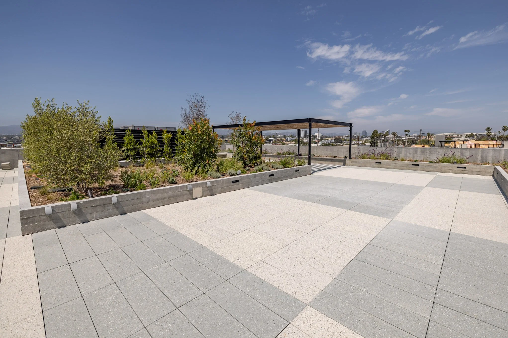 Rooftop terrace with outdoor garden beds, paved flooring, a black pergola, and a city skyline in the background under a partly cloudy sky.