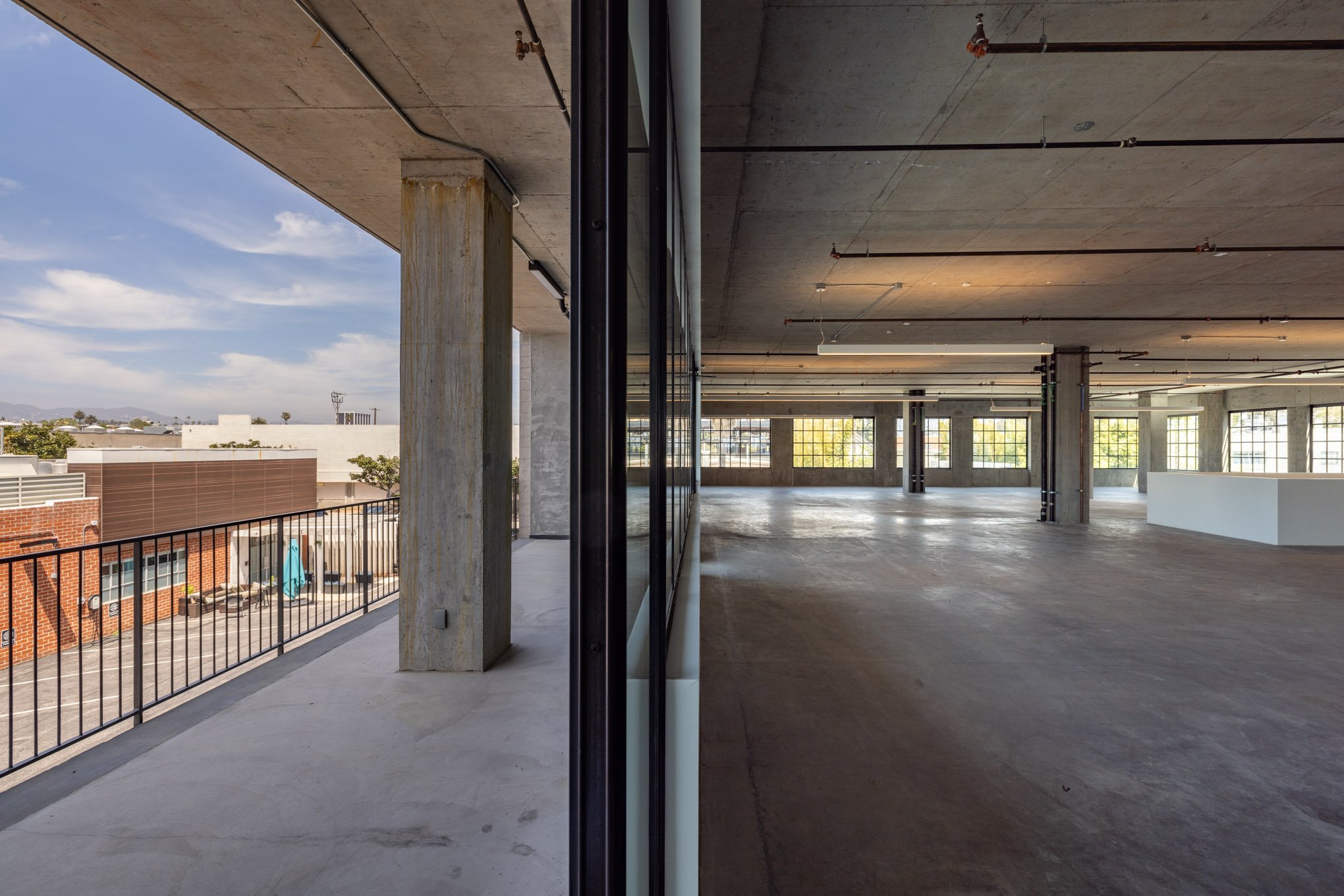 Empty, unfinished interior space with large windows, concrete floors and pillars, and ceiling pipes, viewed from a wall with a glass door.