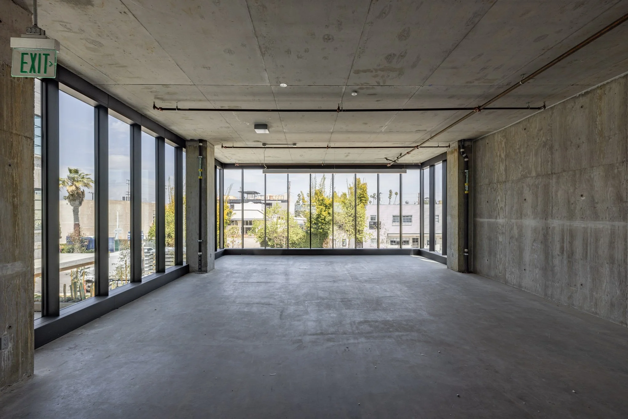Empty unfinished room with large windows, concrete floor and walls, and construction exposed piping, with an exit sign in the corner.