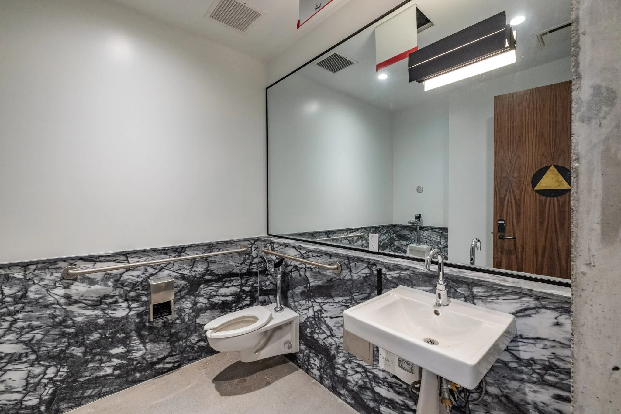 Public restroom with marble walls, a wall-mounted sink, a wall-mounted toilet, a large mirror, and wooden door in the background.