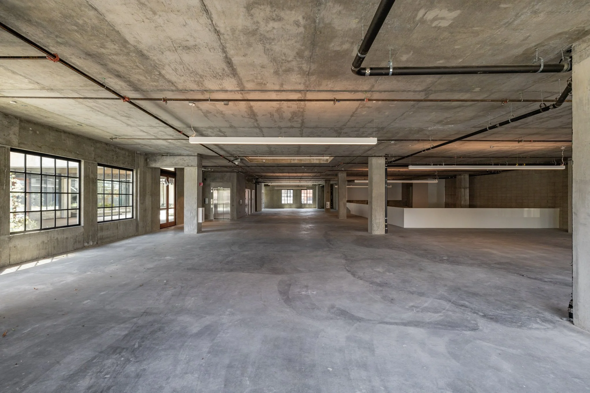 Empty, unfinished commercial building interior with concrete floors, walls, and ceiling, large windows, and exposed piping and lighting fixtures.