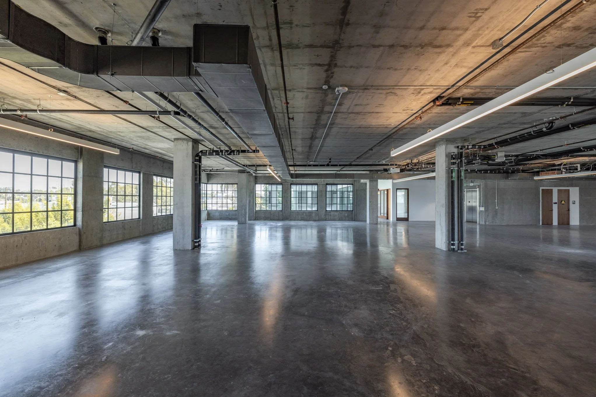 Empty modern industrial-style interior space with concrete floors, large windows, and exposed ceiling ductwork and wiring.