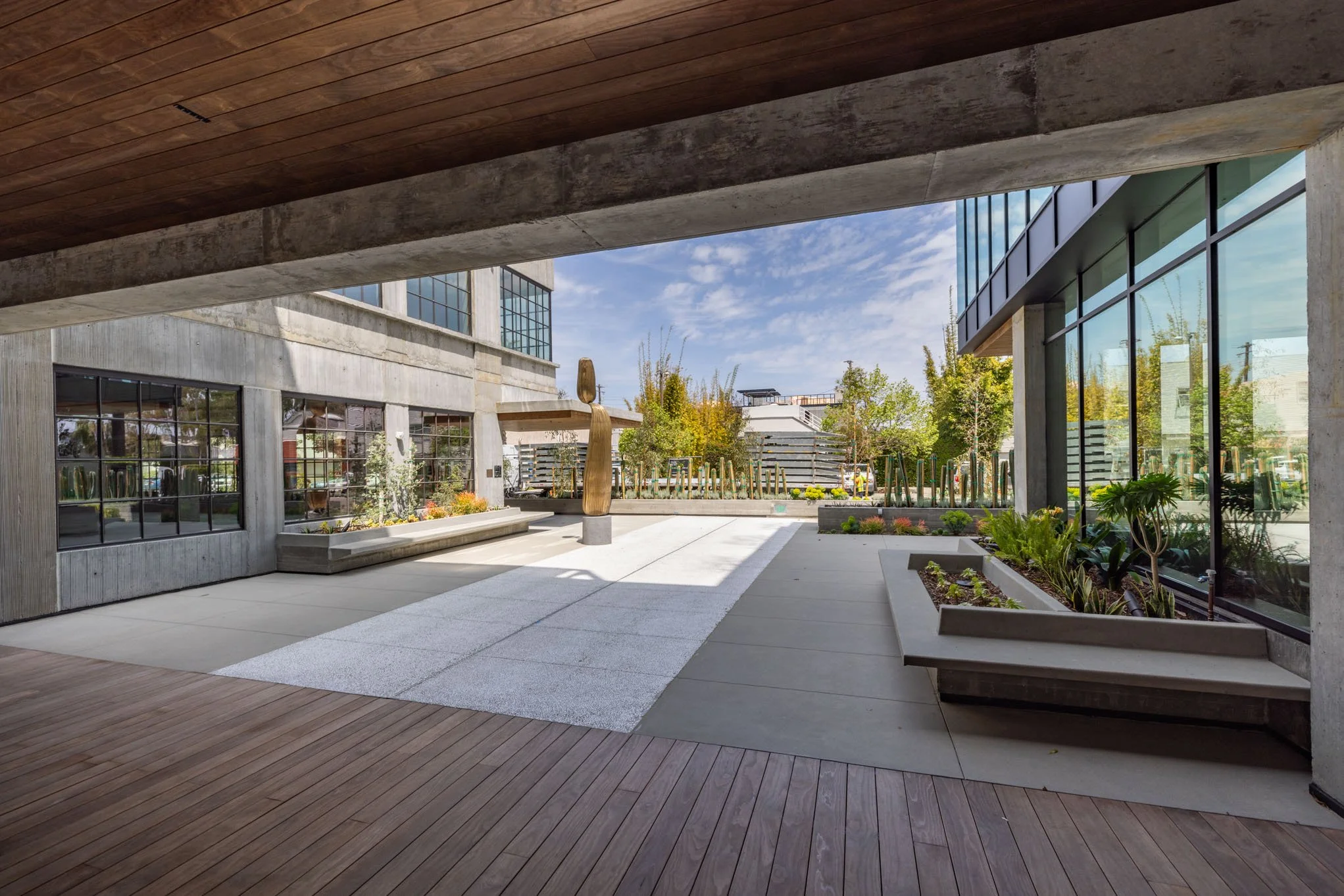Modern outdoor patio area with wooden and concrete flooring, large glass windows, and a landscaped garden with trees and plants, under a wooden overhang.