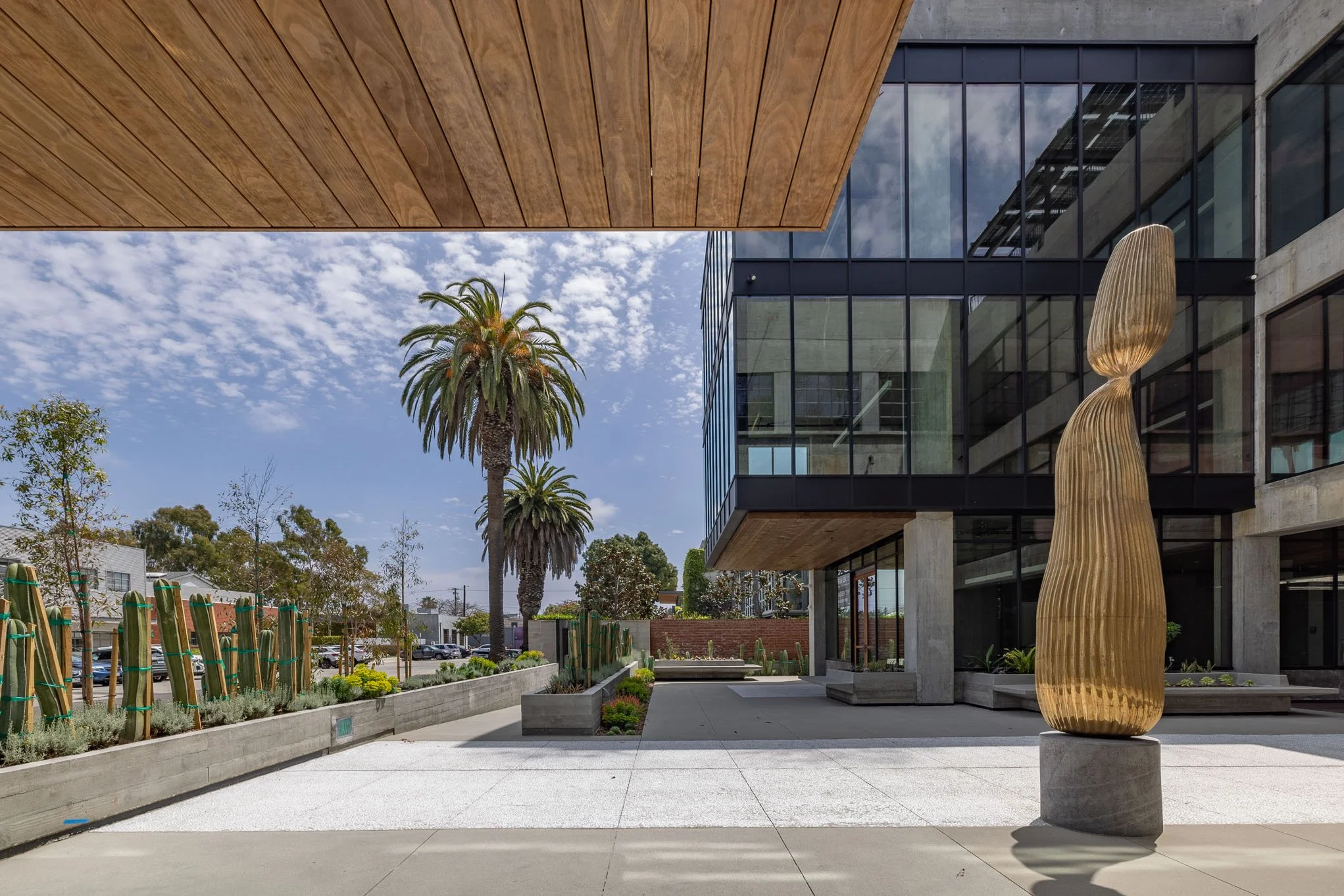 Modern building entrance with glass windows, a wooden overhang, and a large gold sculpture outside, with palm trees and cacti in the landscaping.