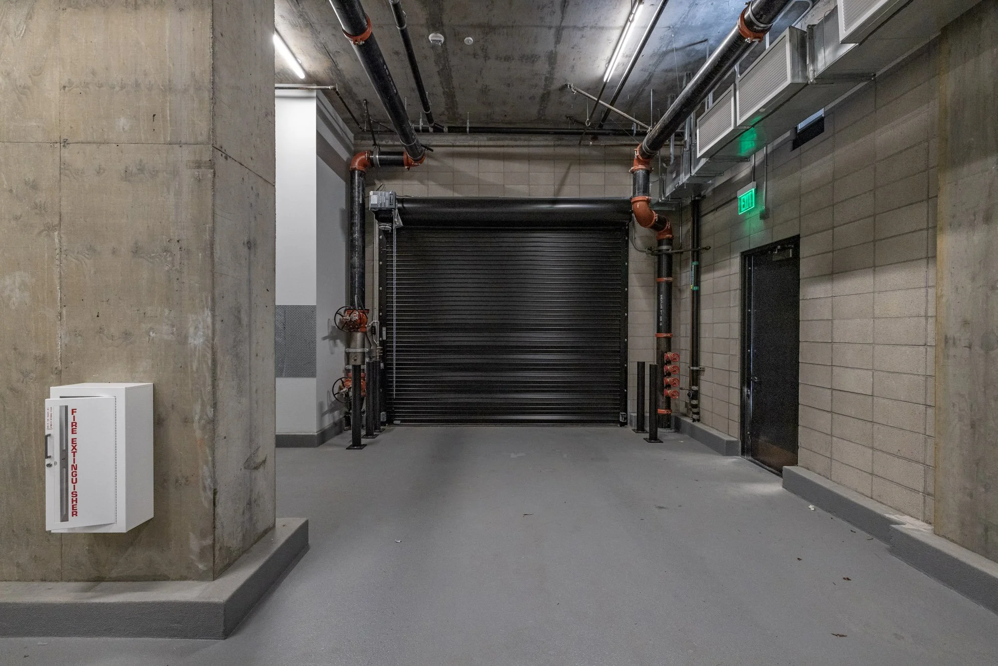 Underground garage or maintenance area with a closed black rolling shutter door, exposed pipes, green exit signs, and a fire extinguisher cabinet on a concrete wall.