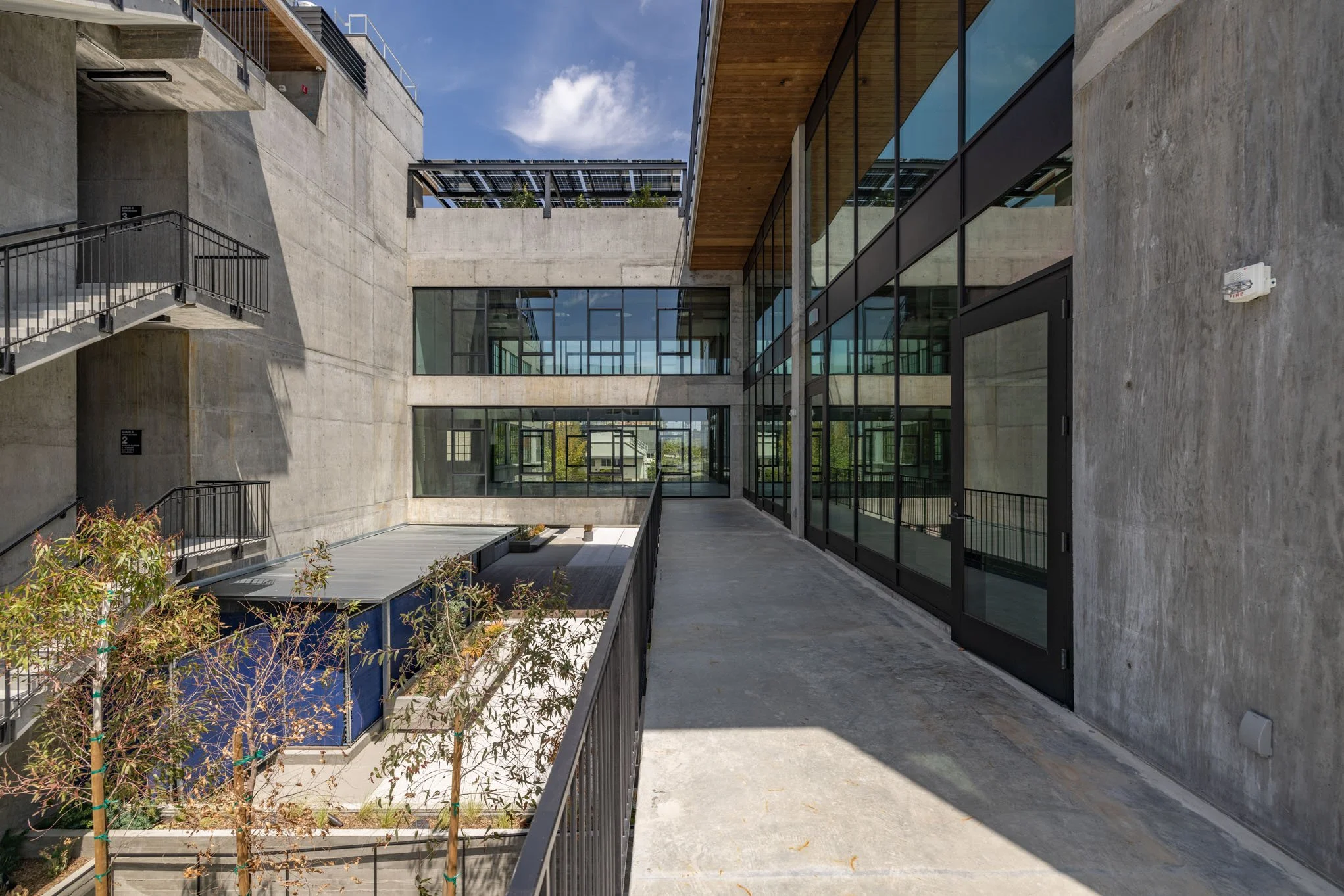 Modern building exterior with concrete walls, large glass windows and doors, outdoor corridor, metal staircases, and some small trees