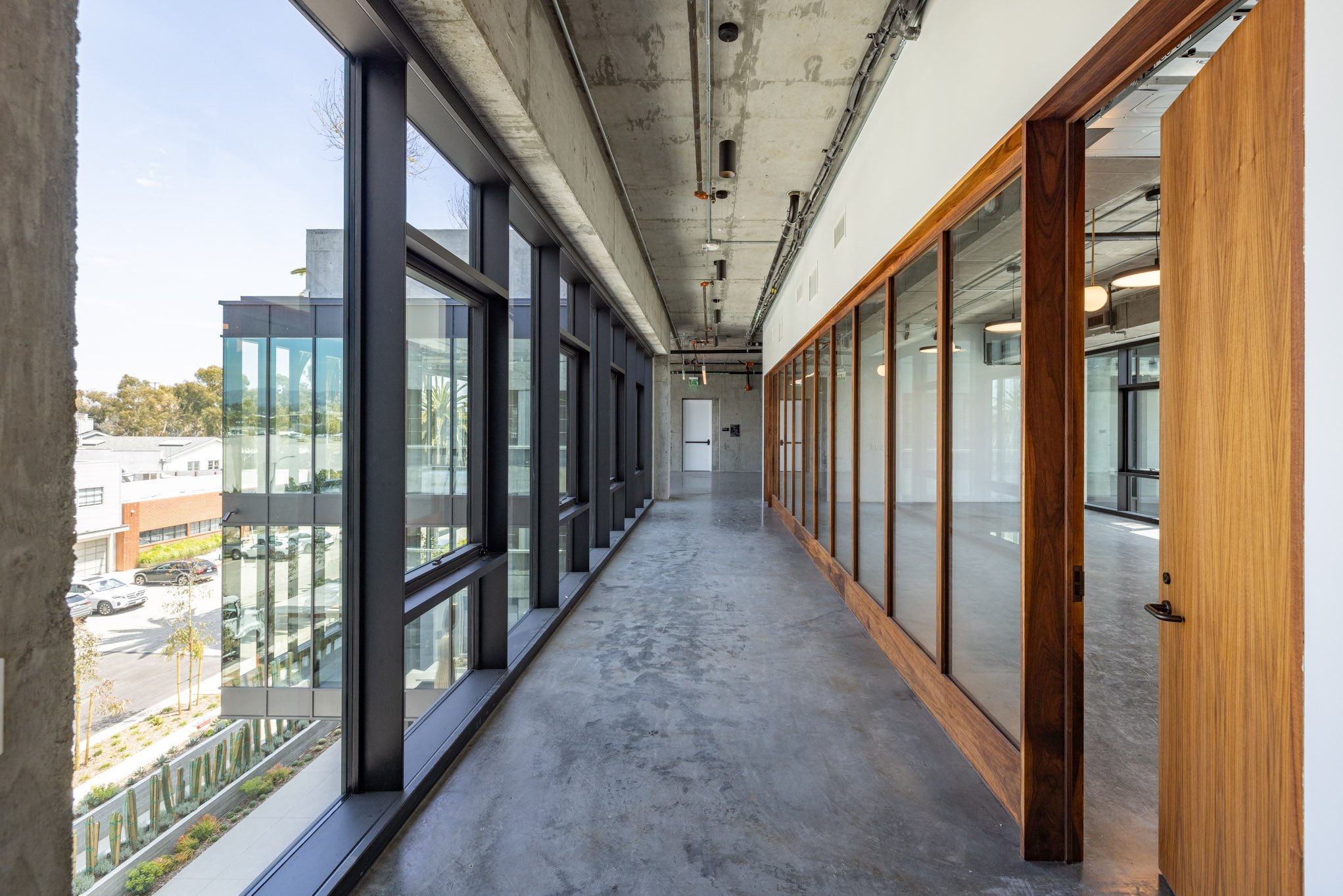 Modern interior corridor with large floor-to-ceiling windows on the left and a glass partition with a wooden door on the right.