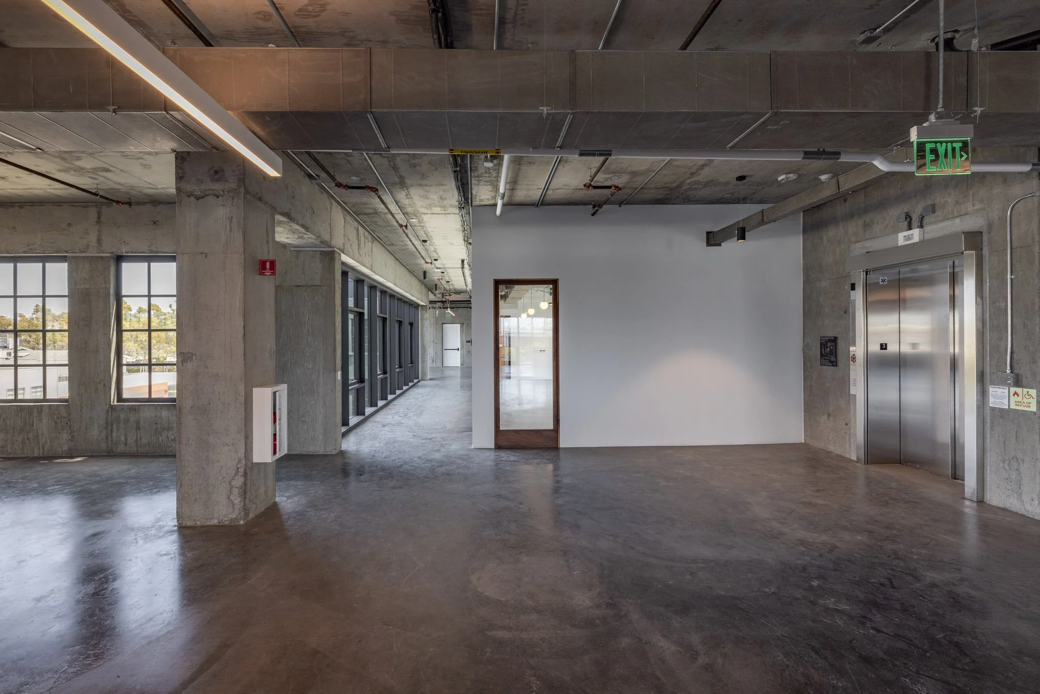 Empty modern industrial lobby with elevator, concrete walls and floors, large windows, and green exit sign.