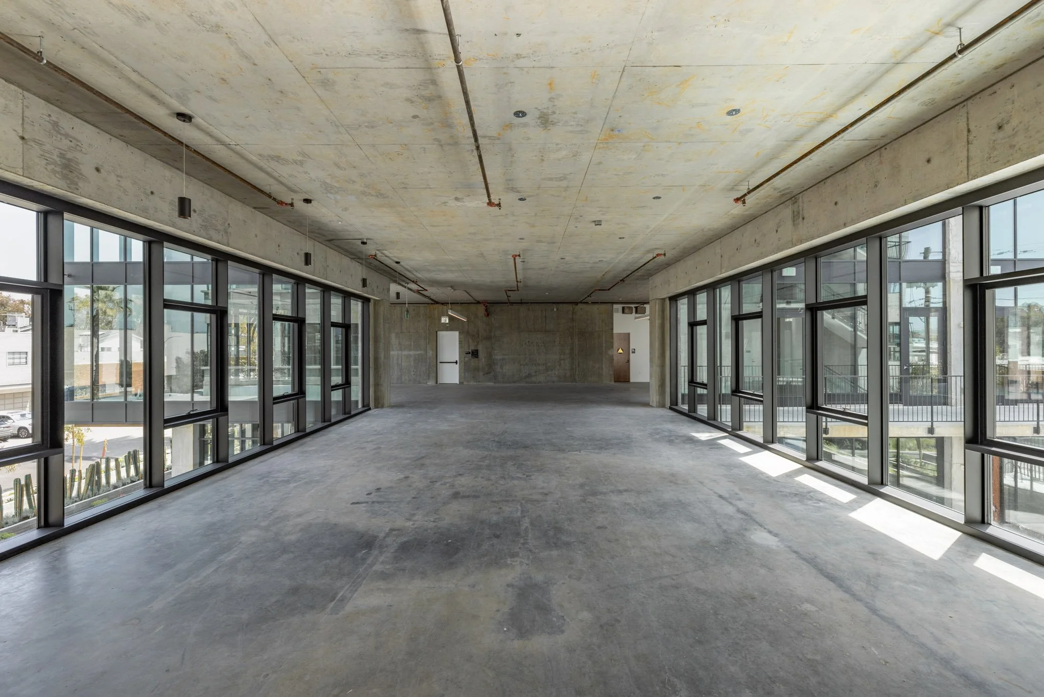 An empty, unfinished commercial space with large floor-to-ceiling windows on both sides, exposed concrete walls and ceiling, and a concrete floor.