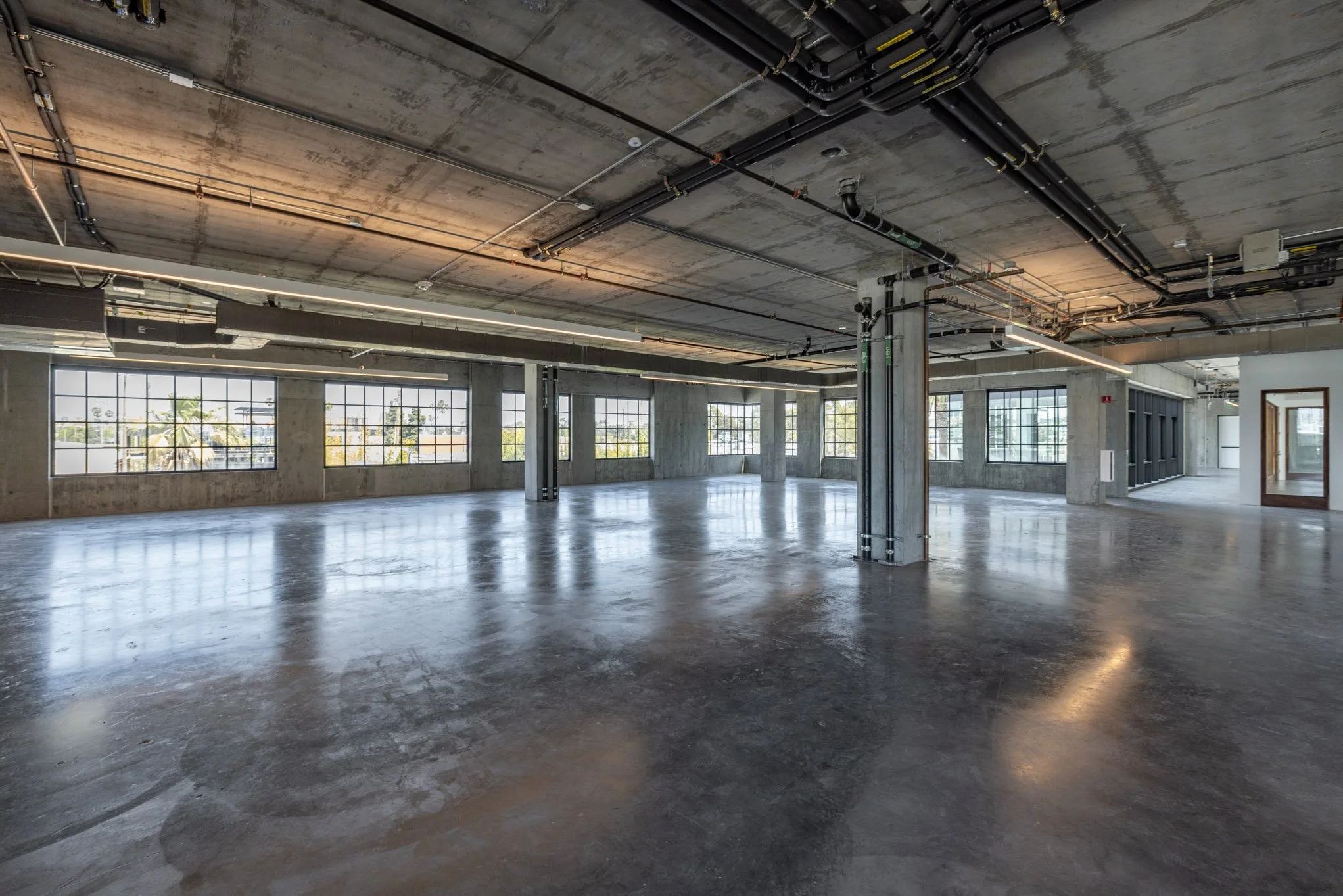 Empty commercial or office space with large windows, concrete floors, exposed ceiling pipes, and structural columns.