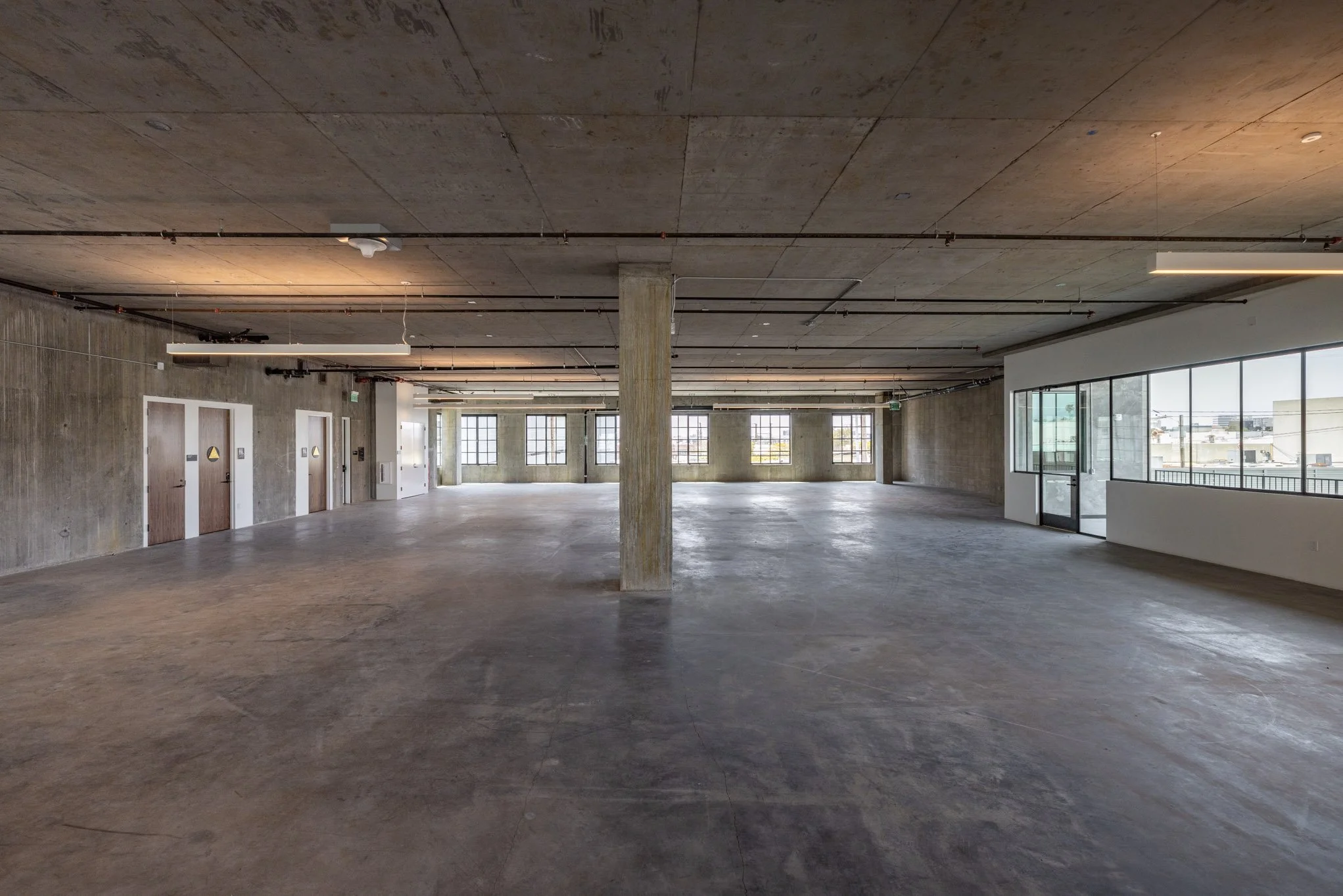 Empty industrial-style interior space with large windows, concrete floors, exposed ceiling pipes, and a structural column in the center.