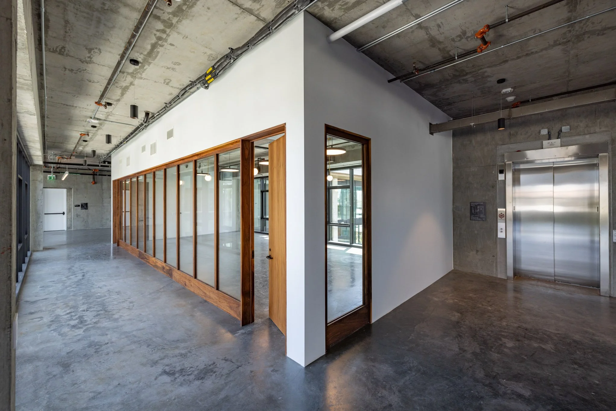 Interior space with concrete floors, exposed ceilings with pipes, a glass-enclosed room with wooden frames, and an elevator with metal doors.