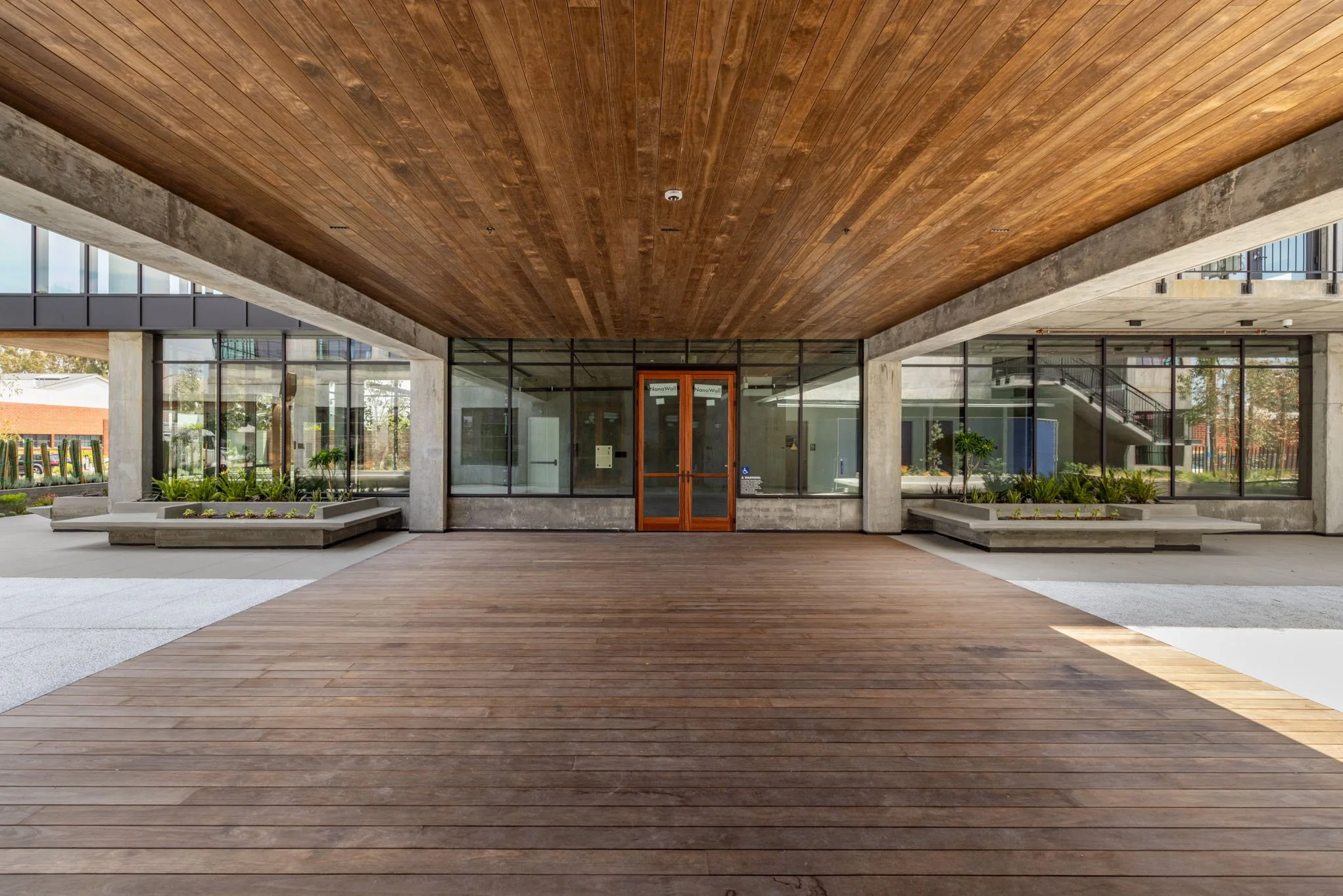 Modern building entrance with large glass windows, wooden and concrete architectural elements, and landscaped planters.