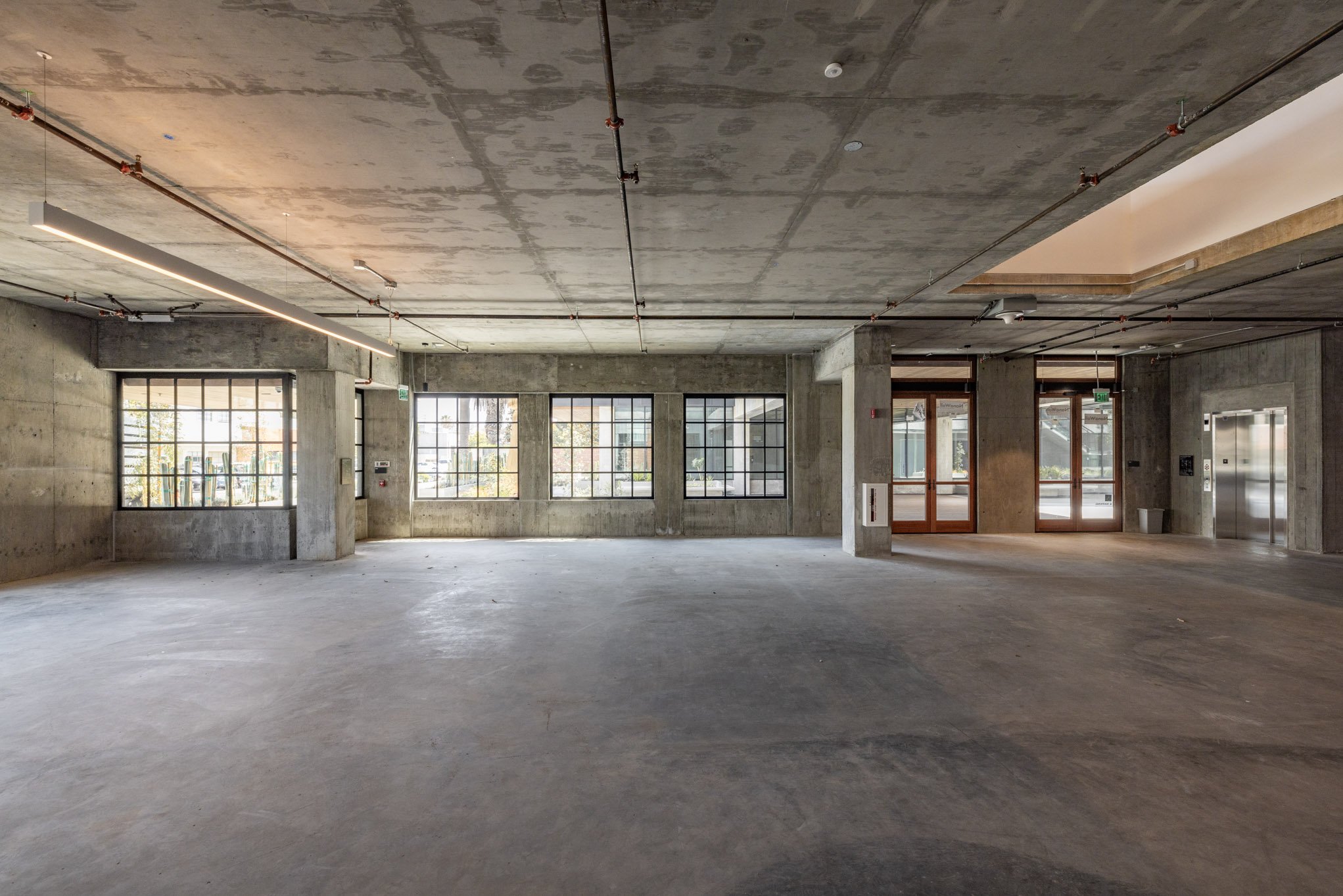 Empty industrial-style interior with concrete floors, concrete walls, large windows, and an elevator on the right. Exposed piping and ceiling details