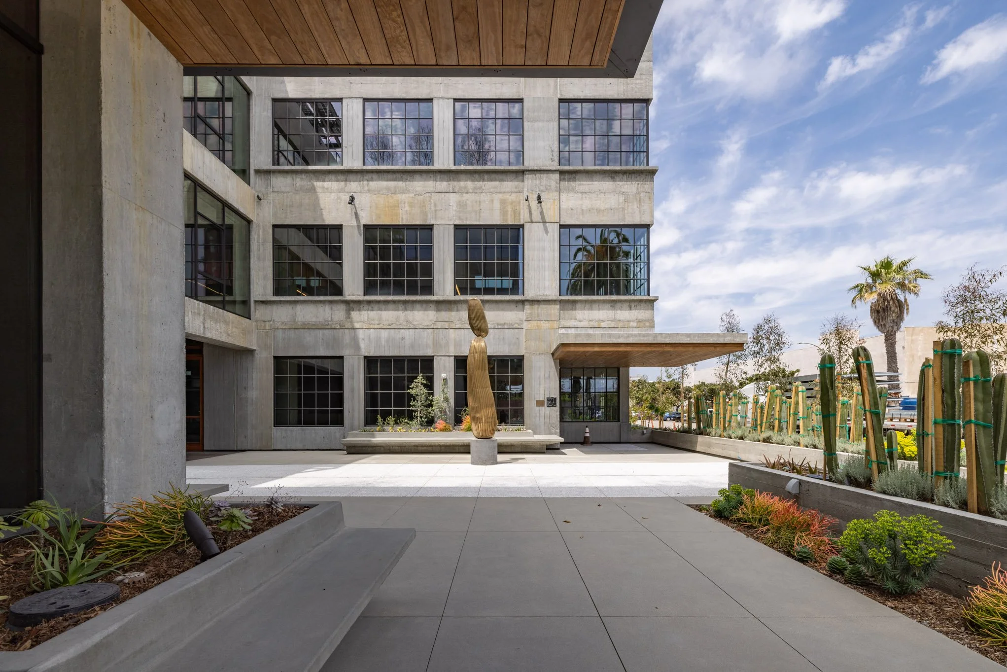 Modern multi-story building with large grid windows, a small garden area with cacti on the right, and a sculpture resembling a gold stacked figure in the center of the courtyard. The sky is partly cloudy.