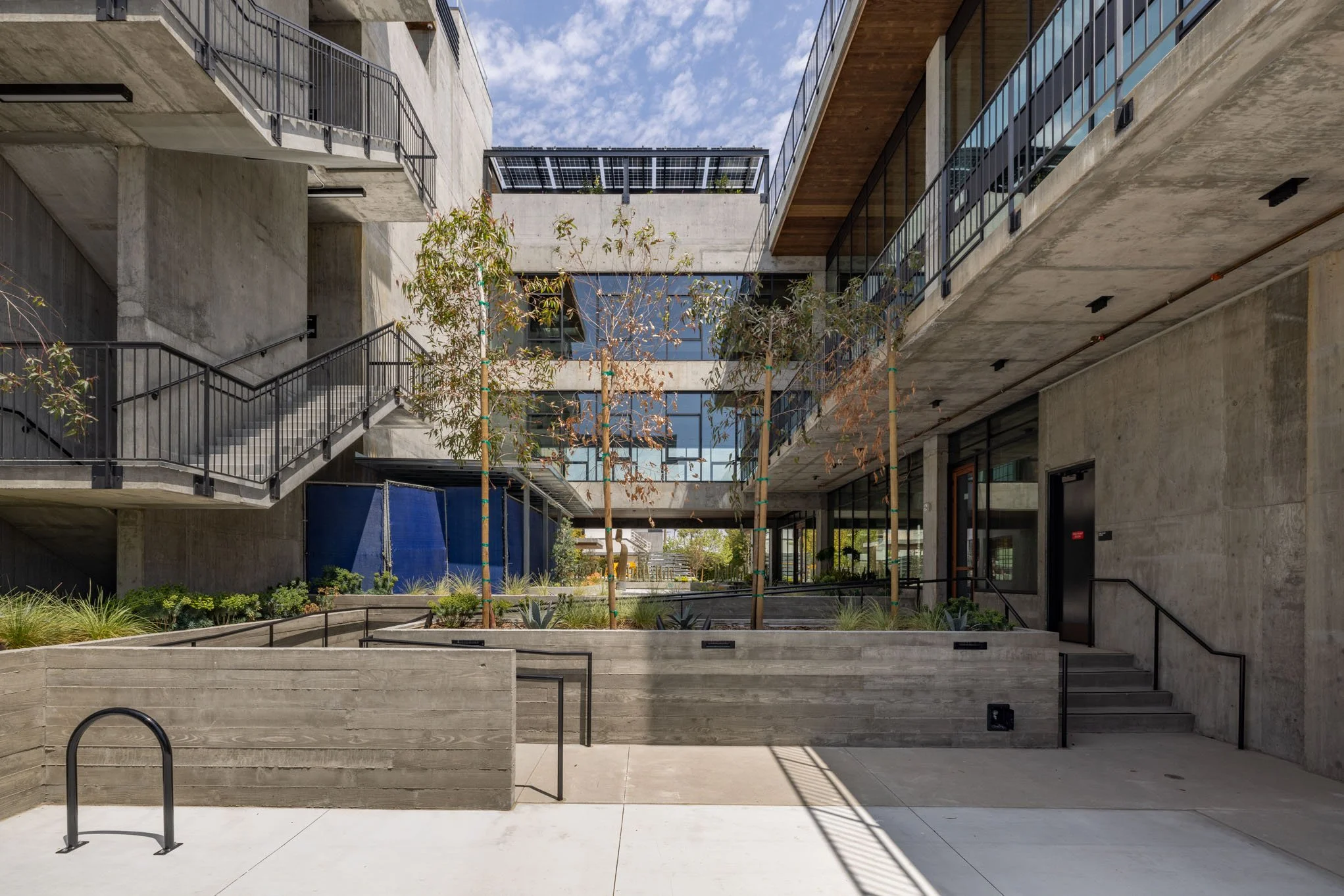 Modern multi-story concrete building courtyard with trees, plants, and stairs, featuring glass railings and solar panels on the roof.