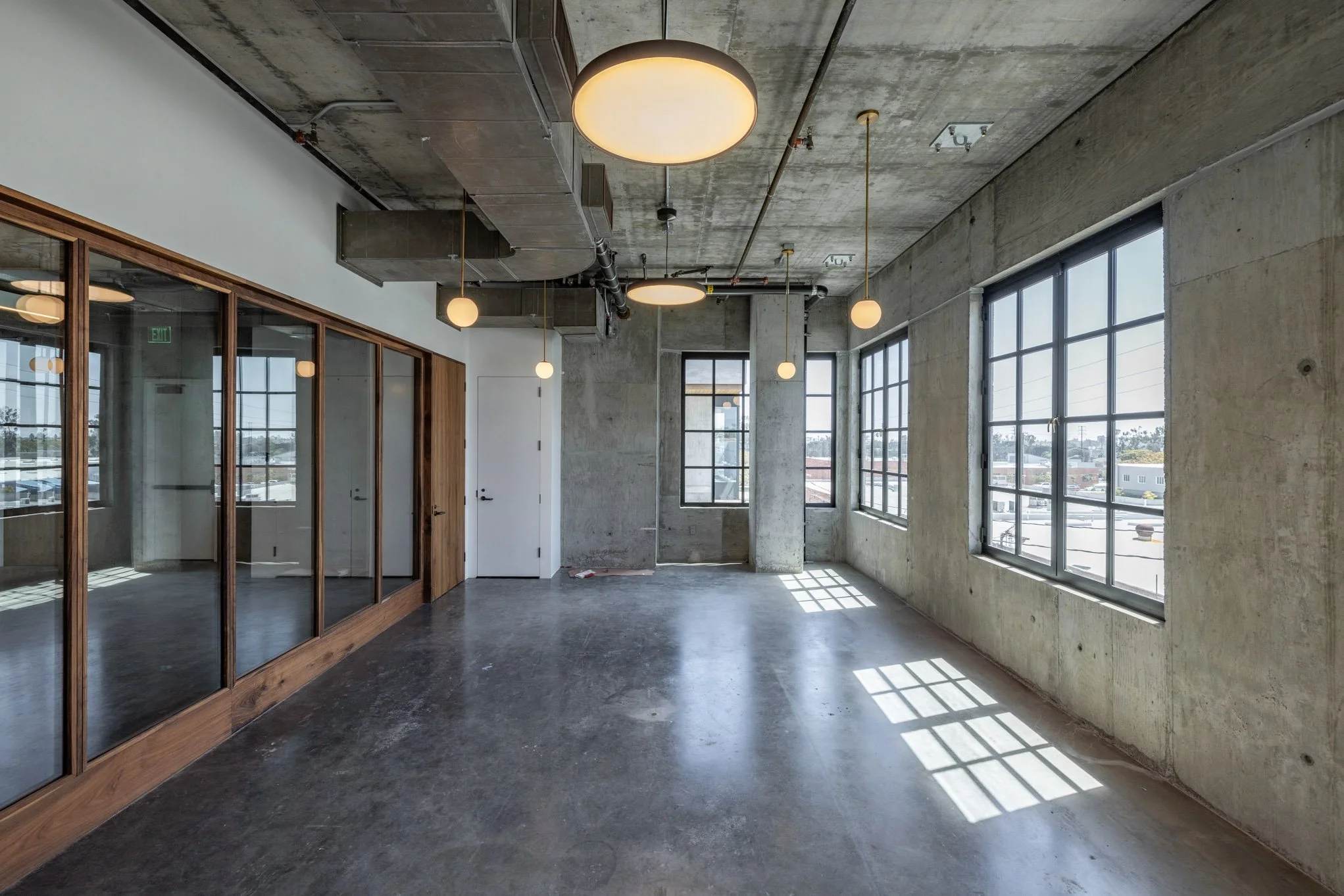 Empty industrial-style room with large windows, exposed concrete walls and ceiling, wooden-framed glass partition, and modern round pendant lights, with sunlight casting grid-patterned shadows on the floor.
