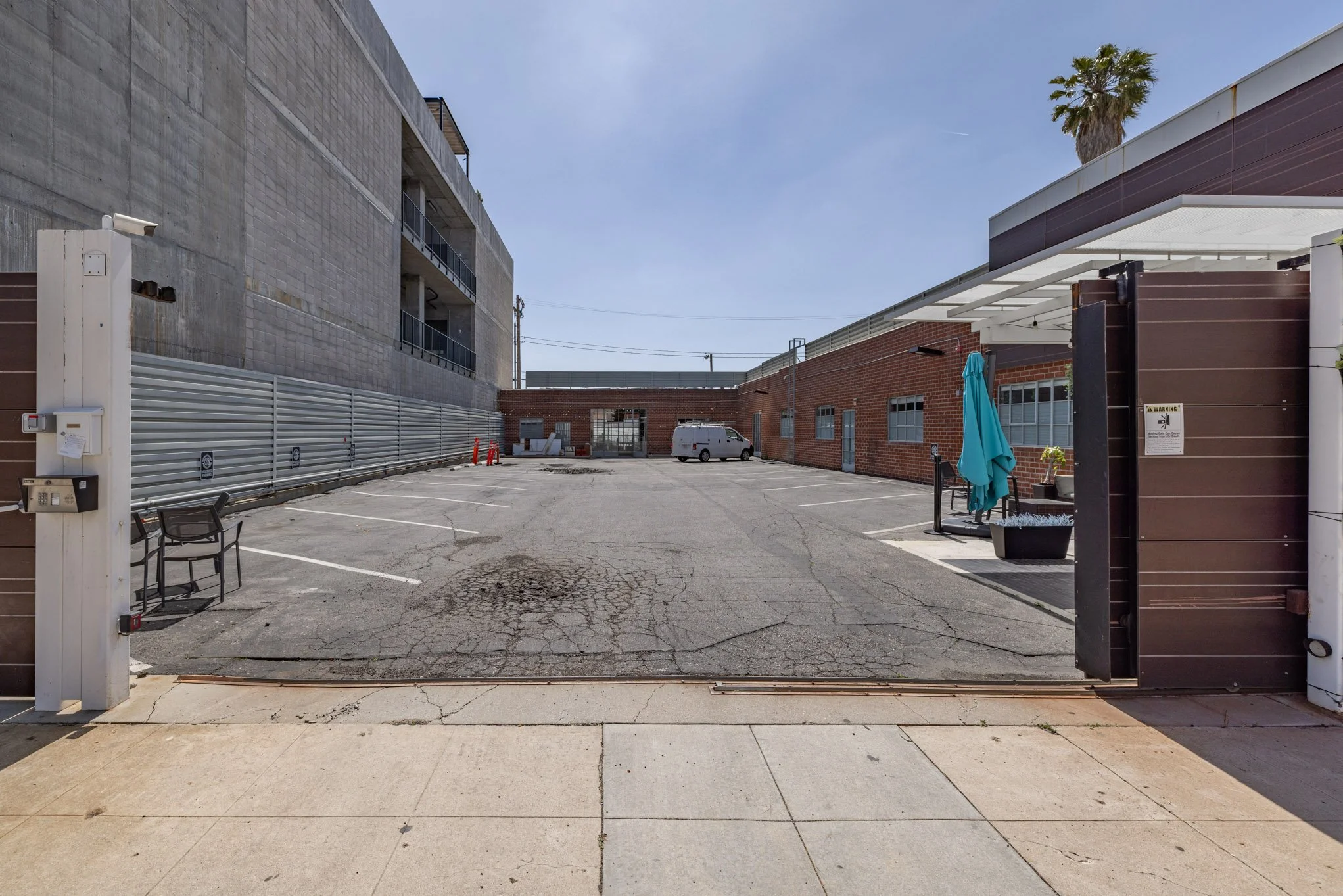Empty parking lot with cracked asphalt, surrounded by a brick building on the right, a multi-story concrete building on the left, shaded seating with an umbrella, and a white van parked in the distance under a clear blue sky.