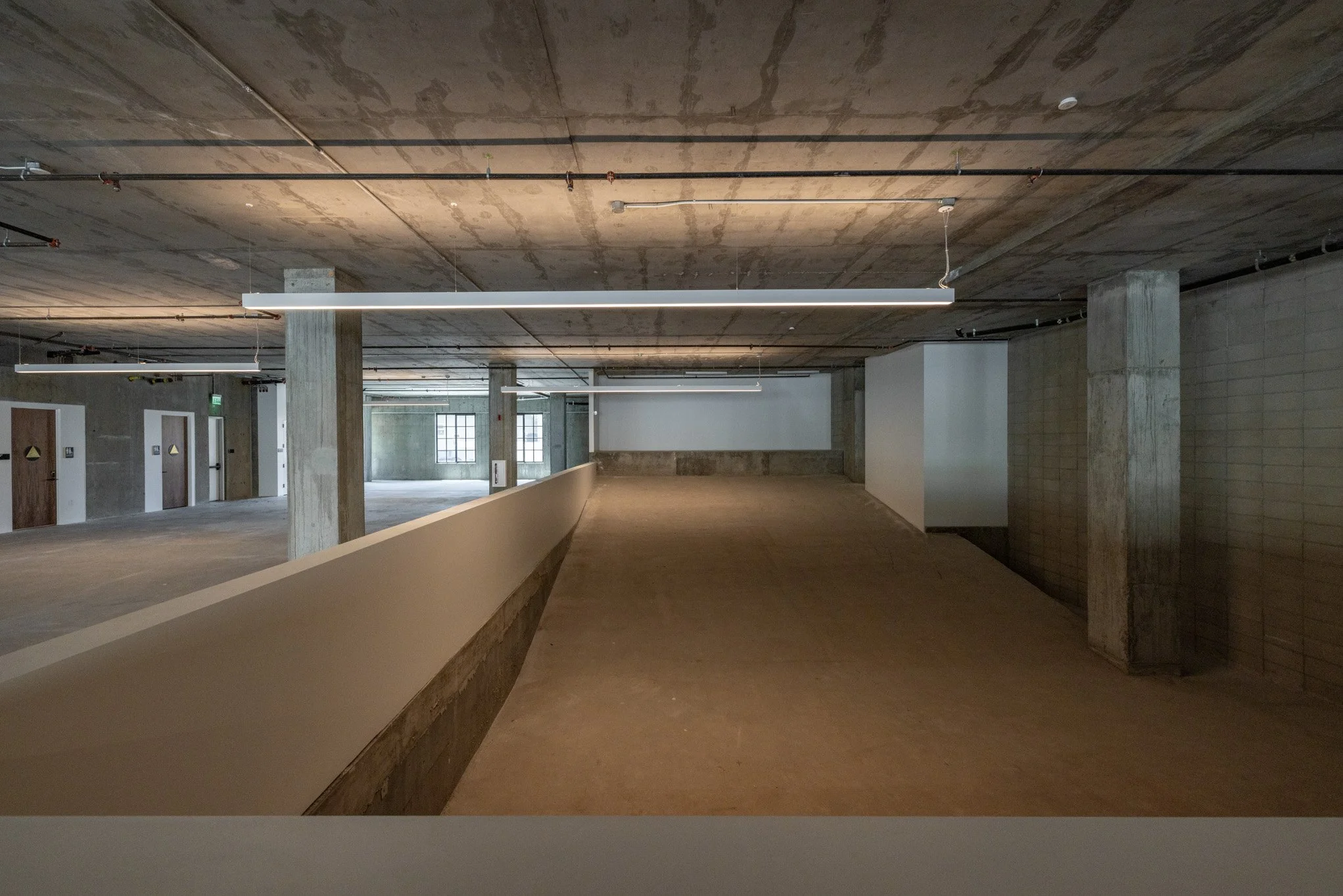 Inside a modern, multi-level parking garage with concrete pillars, exposed ceiling, hanging linear lighting, and a ramp leading to an upper level.