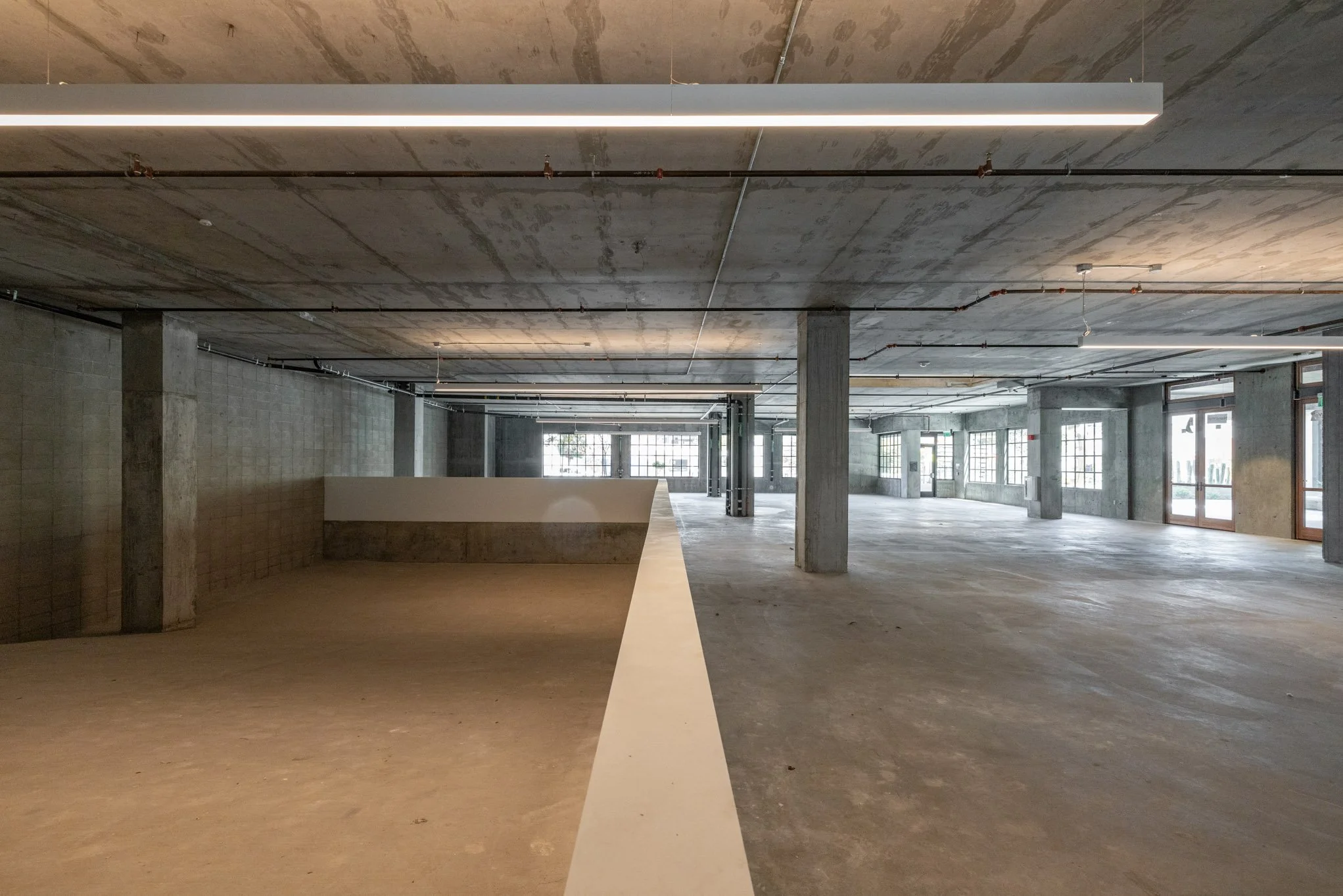 Empty, unfinished commercial space with exposed concrete walls, ceiling, and floor, illuminated by natural light from large windows and glass doors on the right.