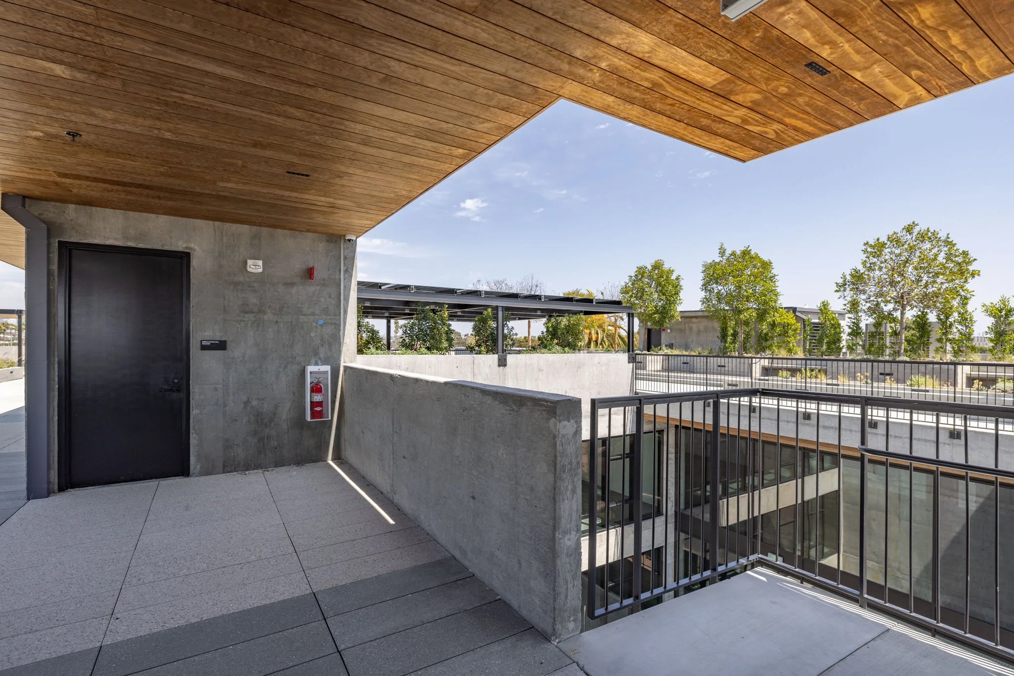 Outdoor balcony area with concrete walls, black door, fire extinguisher, metal railing, trees in the background, and a wooden ceiling.