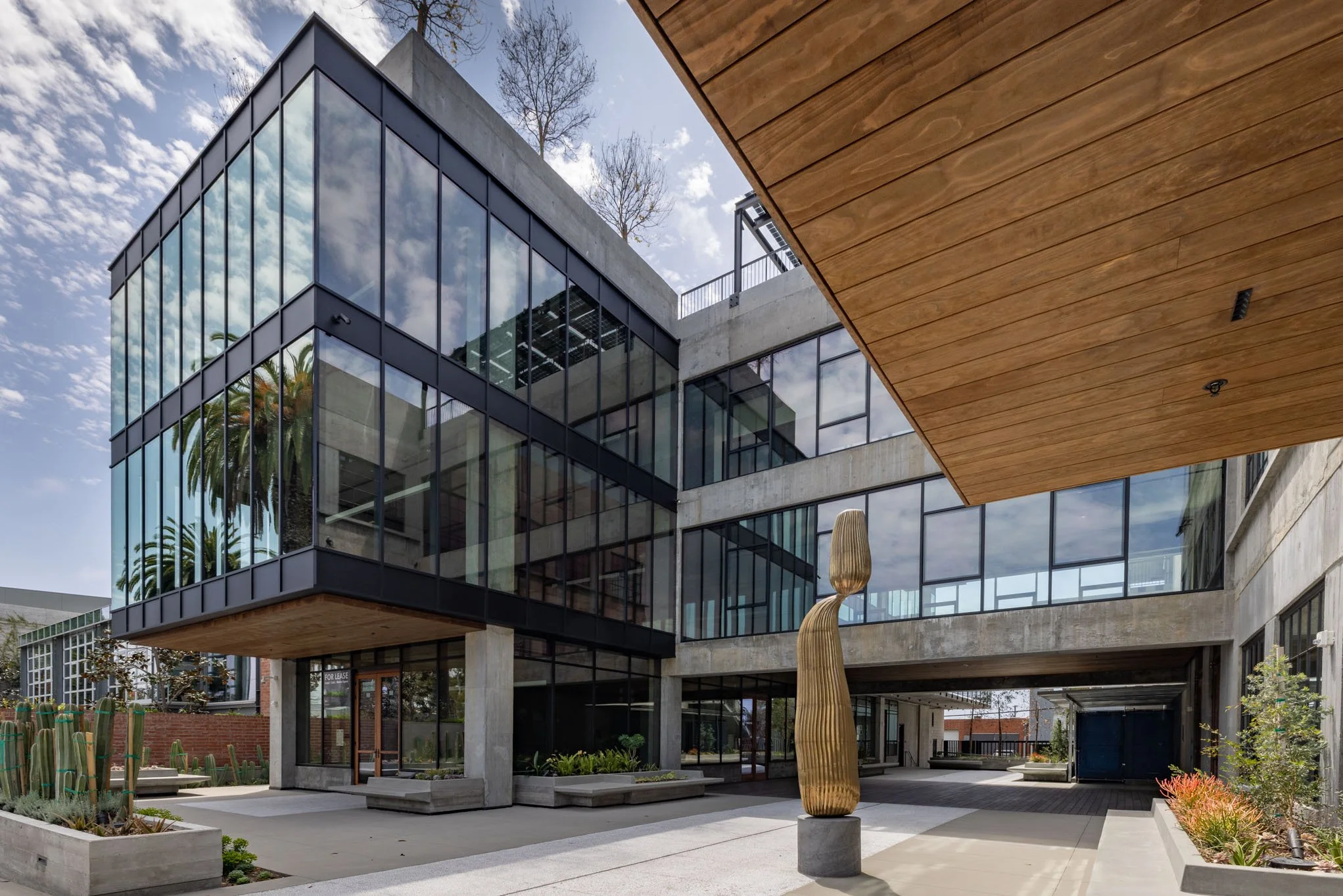 Modern commercial building with large glass windows, concrete accents, and wooden ceiling panels, featuring outdoor landscaping with cactus plants, a carved wooden sculpture, and a clear blue sky.
