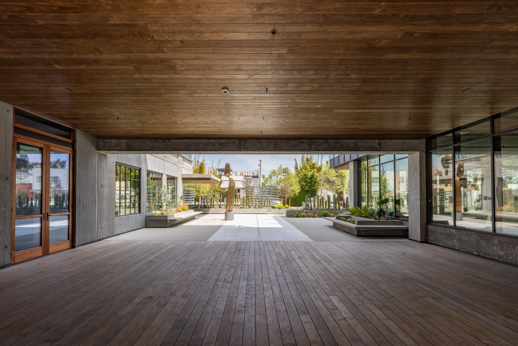 View from inside a covered area looking out onto a courtyard with trees, plants, sculptures, and surrounding modern buildings.