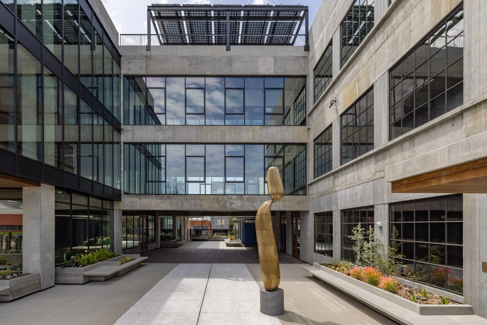 Modern multi-story building with large glass windows, concrete walls, and an outdoor courtyard featuring a wooden sculpture on a concrete base, planters with plants, and benches.