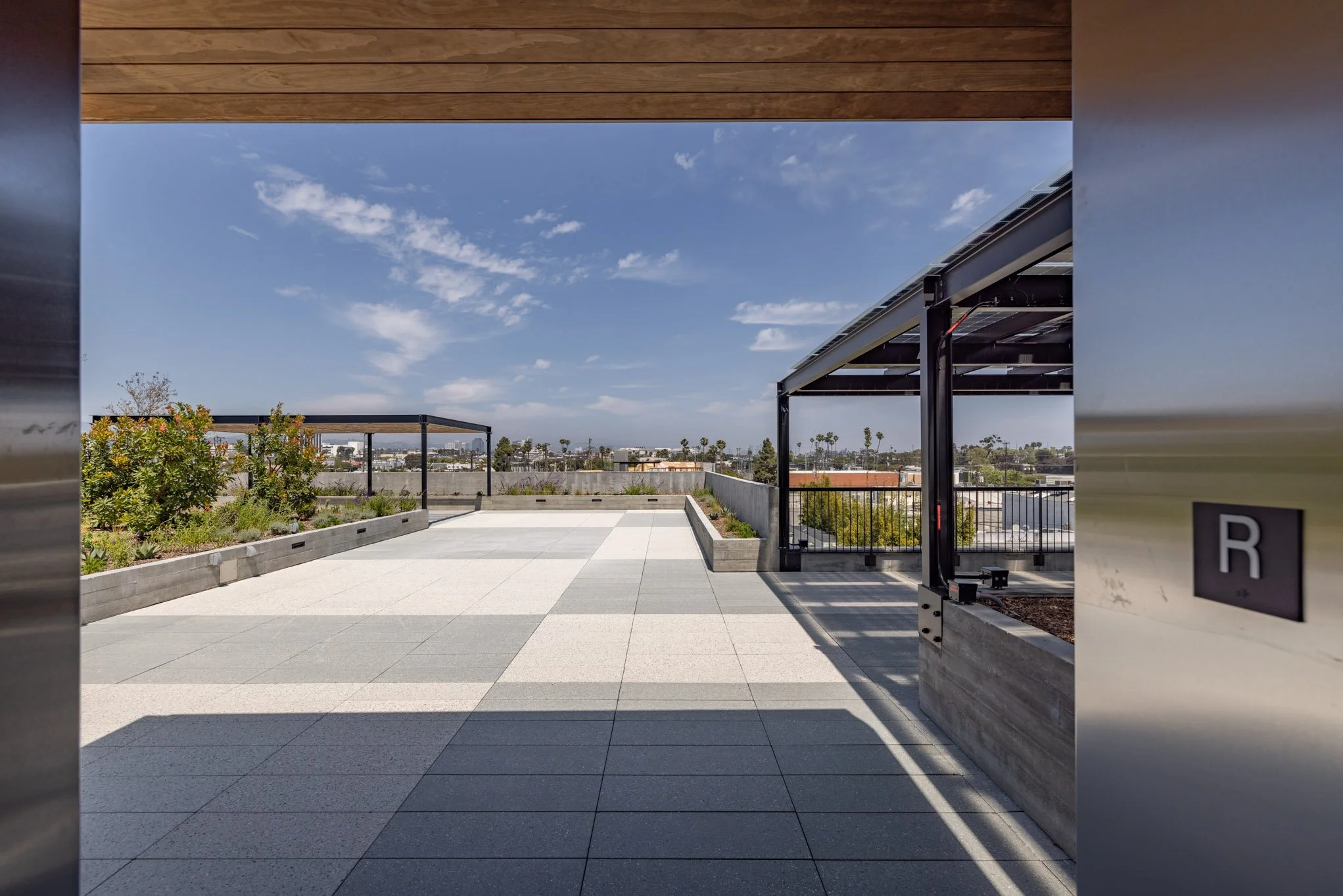 Rooftop terrace with plant bed, modern tile floor, metal pergola, city skyline with palm trees, blue sky with clouds