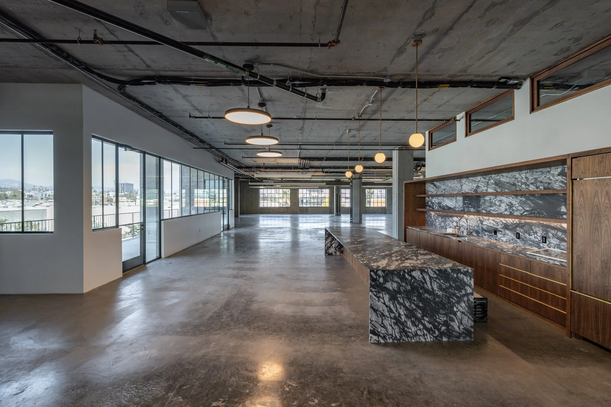 Empty modern office space with large windows, polished concrete floors, exposed ceiling pipes, round ceiling lights, and a marble and wood kitchenette area.