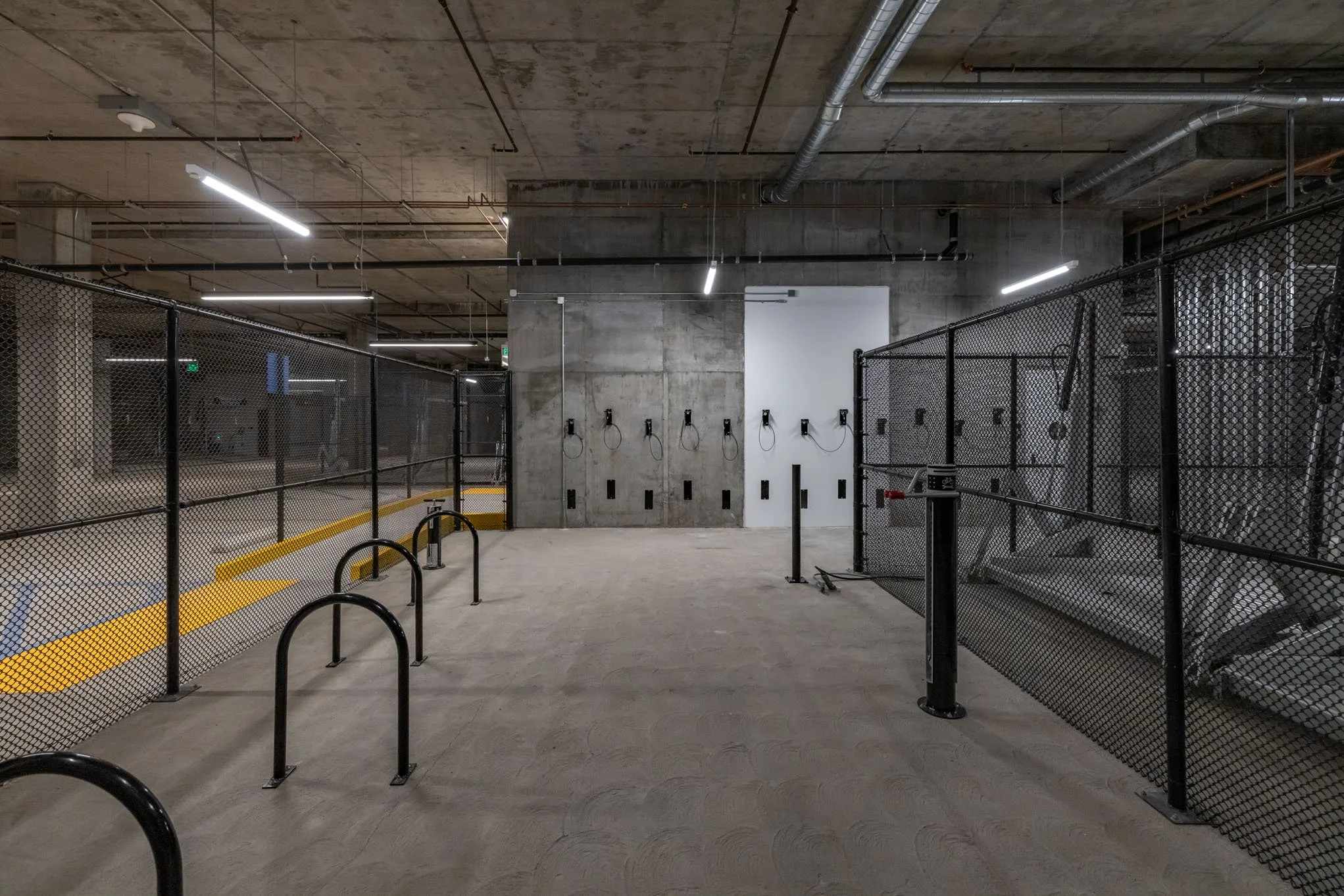 Empty indoor gym space with black safety fences, wall-mounted workout stations, and exposed concrete ceiling and walls.