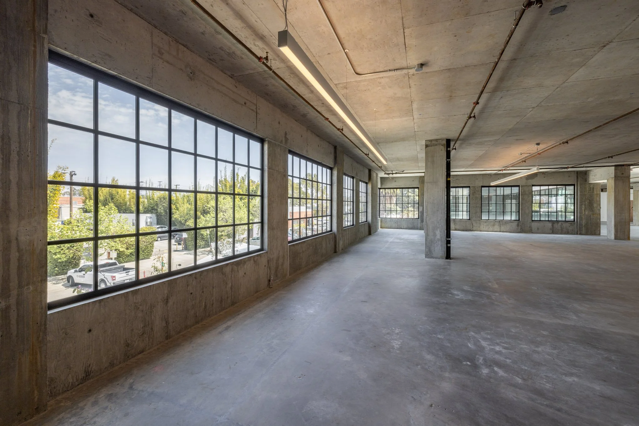 Empty commercial space with large windows, concrete floors, and exposed ceilings with pipes and lighting fixtures.