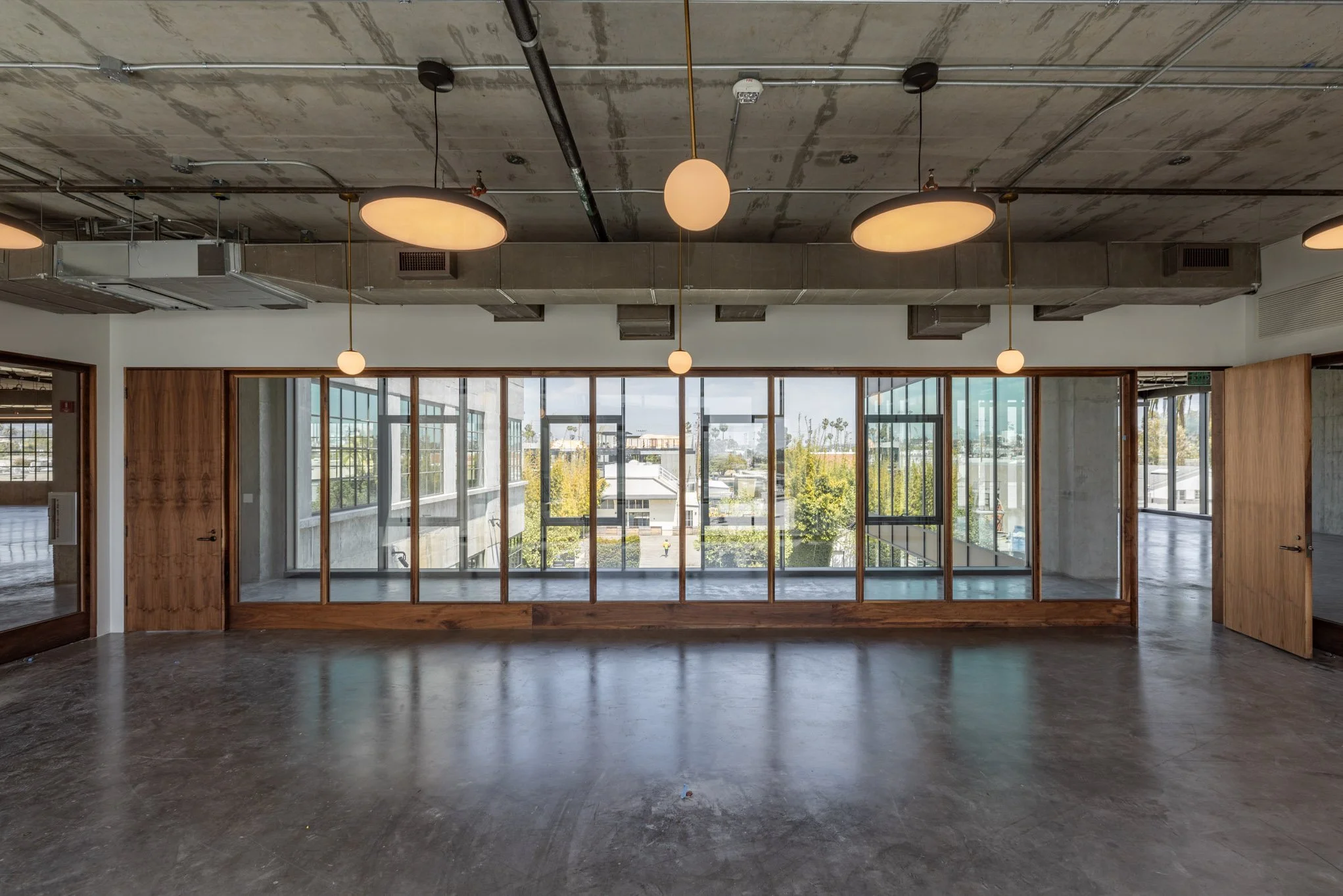 An empty commercial space with polished concrete floors, large windows with wooden framing, and suspended round light fixtures.