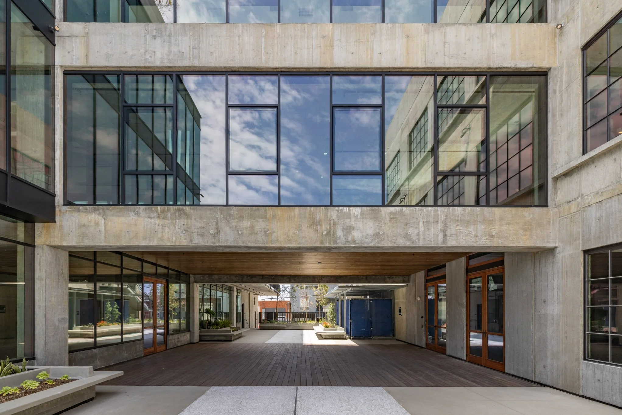 Modern building with large glass windows, concrete facade, and an open ground-level area with wooden flooring and planters.