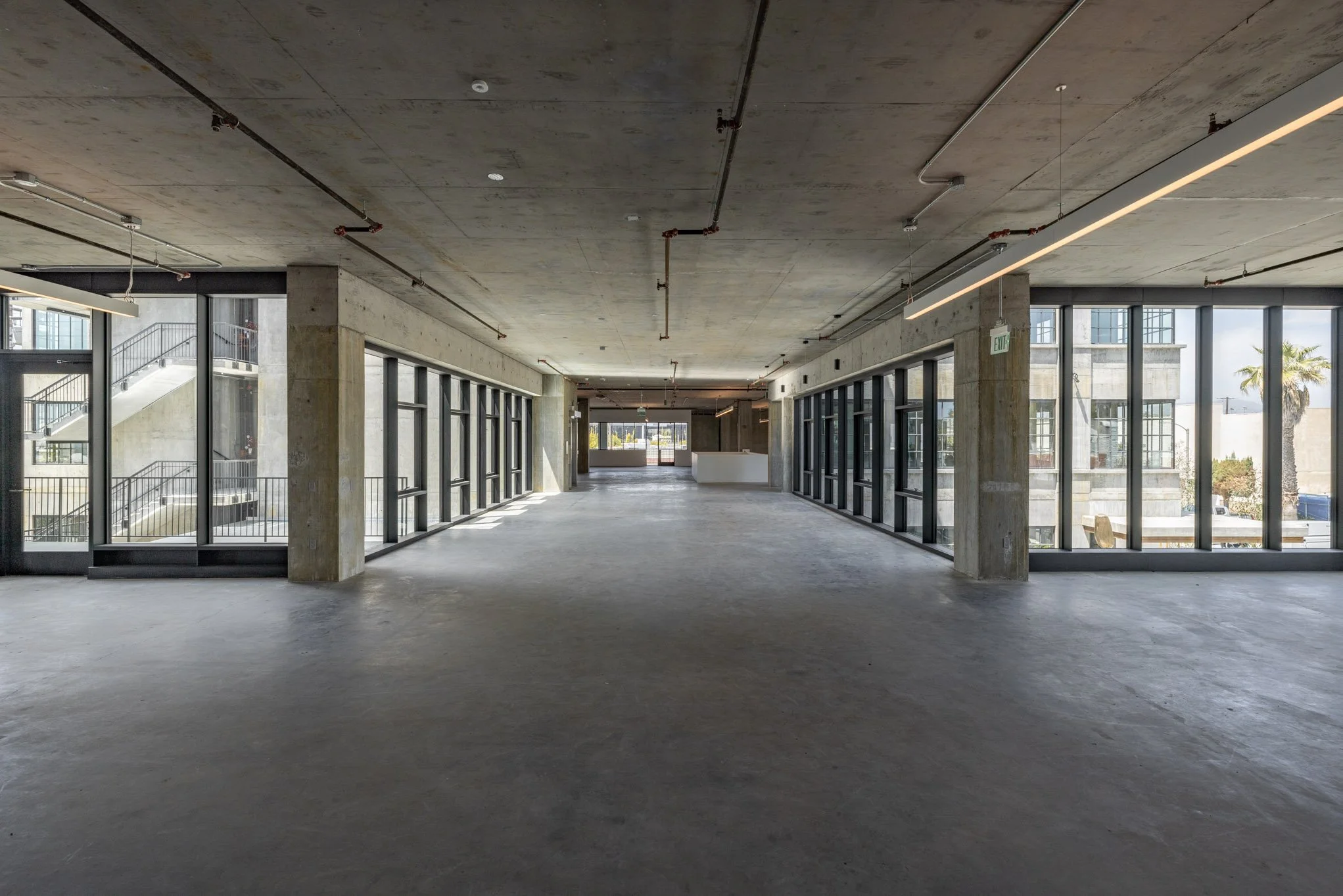 Empty commercial interior space with concrete floors, exposed concrete ceiling with pipes, and large floor-to-ceiling windows on both sides.