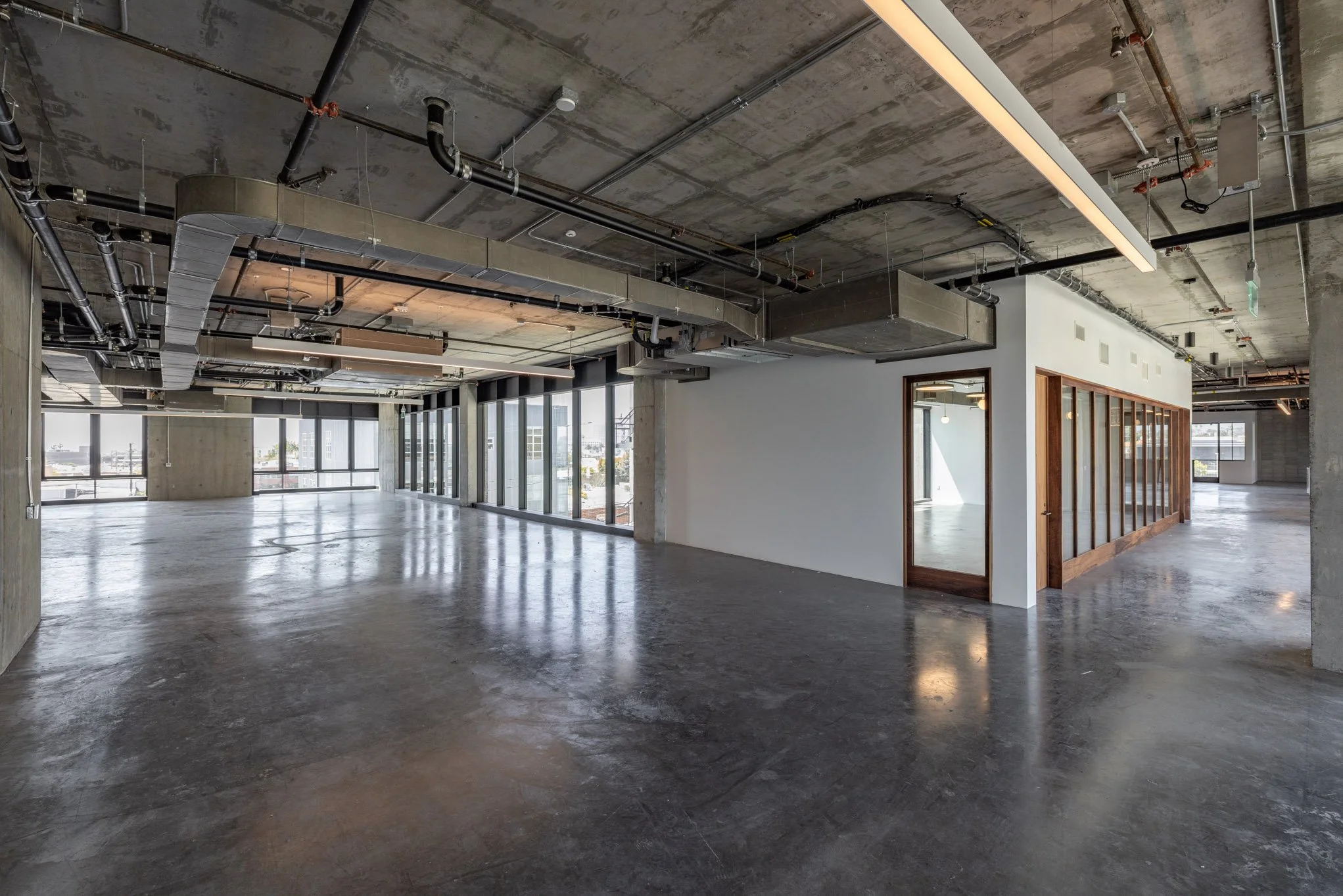 Empty, modern office space with concrete floors, exposed ceiling pipes, large windows, and a small enclosed room with a glass wall and wooden frame.