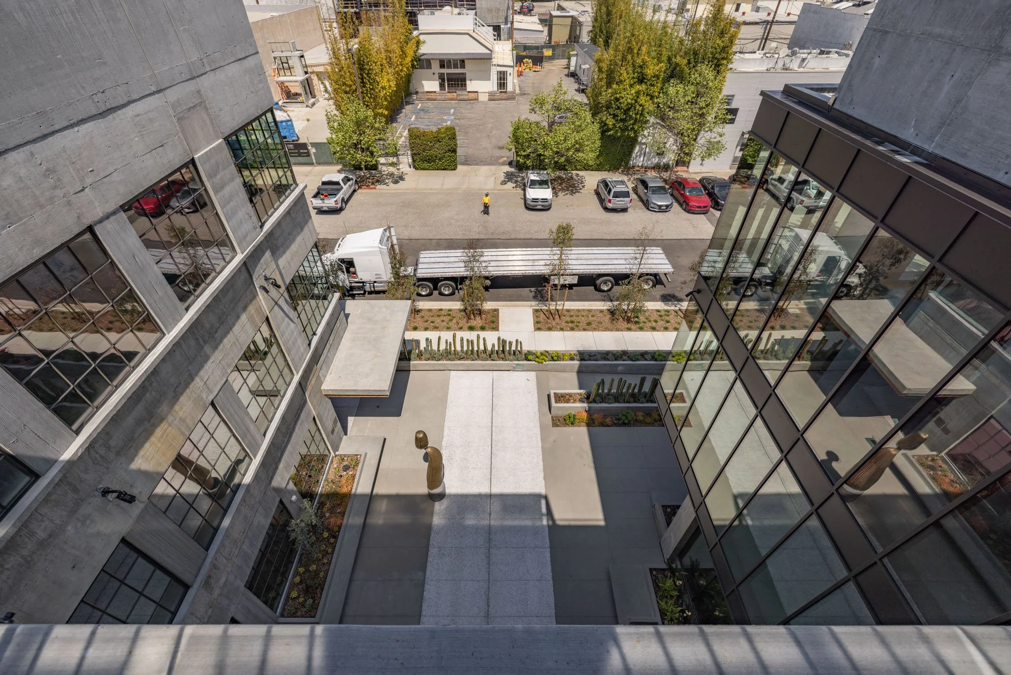 View from a tall building looking down at a courtyard with a sidewalk, small trees, and cactus garden beds, with parked cars and a person walking on the street in the background.