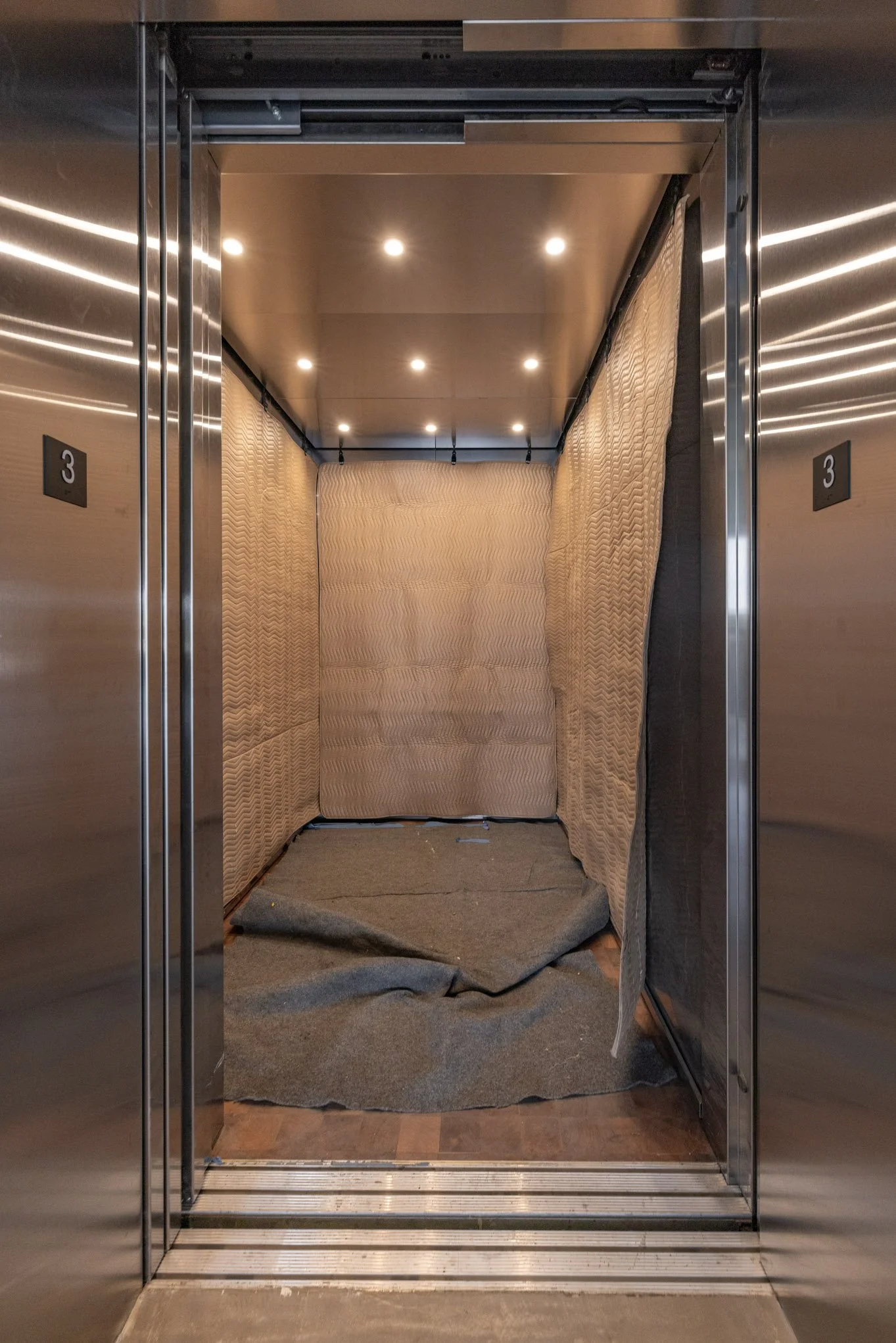 Empty elevator with padded walls and a gray carpeted floor, illuminated by ceiling lights.