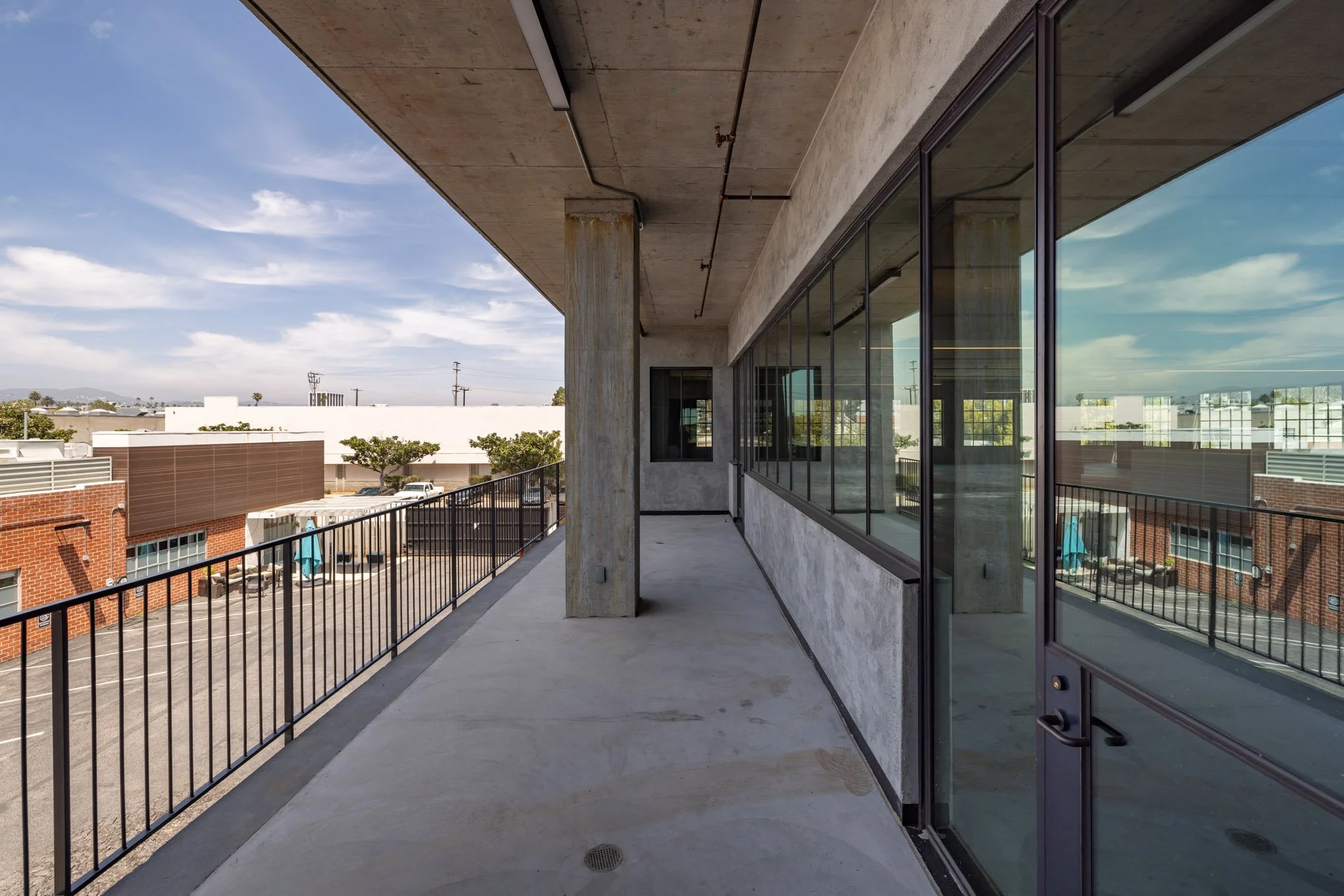 An empty balcony with concrete flooring and black metal railing, overlooking a parking lot and neighboring buildings against a blue sky with clouds.