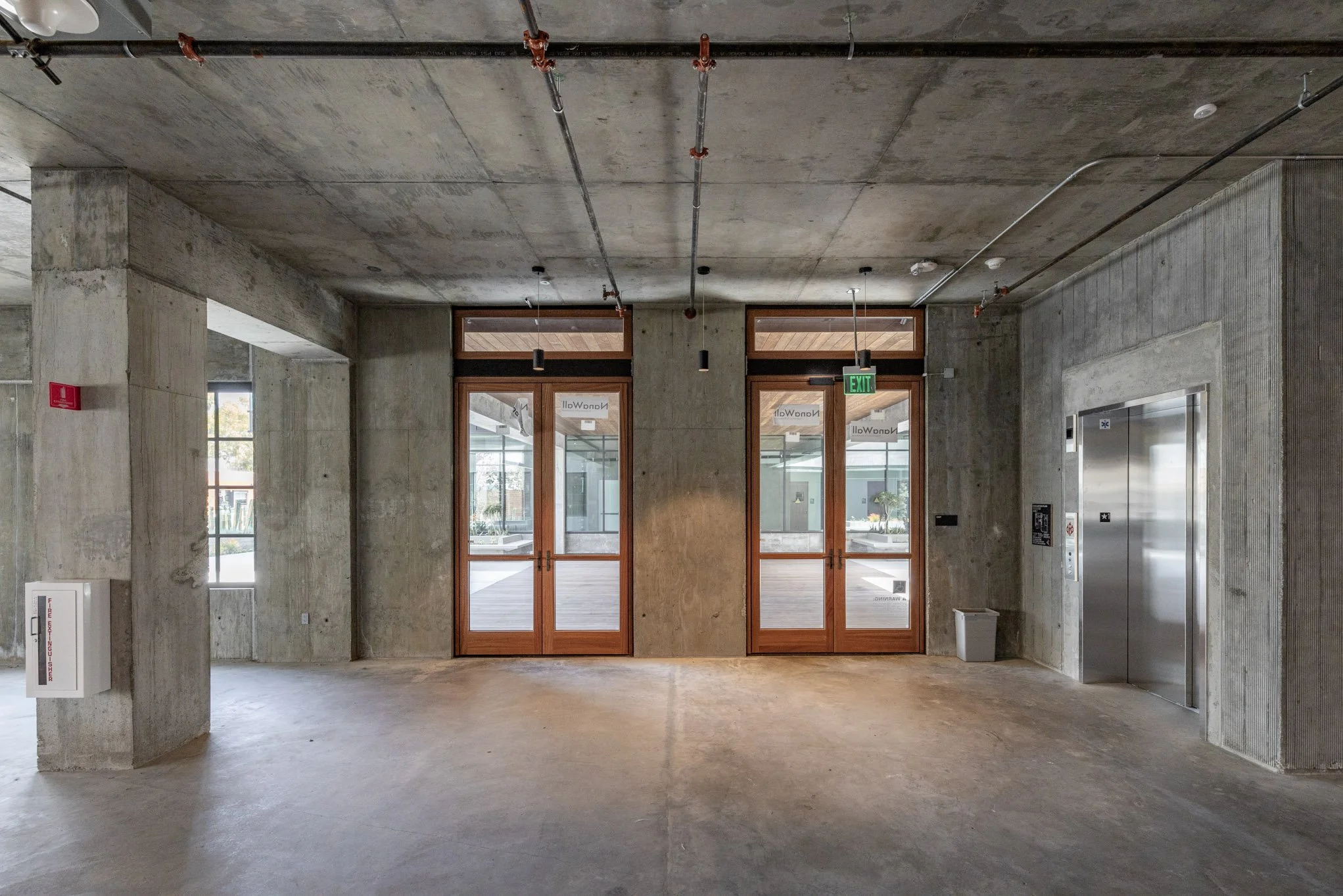 Concrete lobby with glass doors, elevator, exit sign, and fire safety equipment.