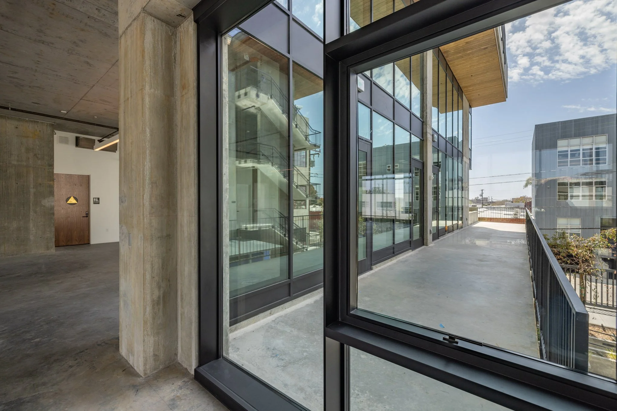 Interior of a modern building with large glass sliding doors leading to a balcony with a black metal railing, overlooking neighboring buildings under a partly cloudy sky.